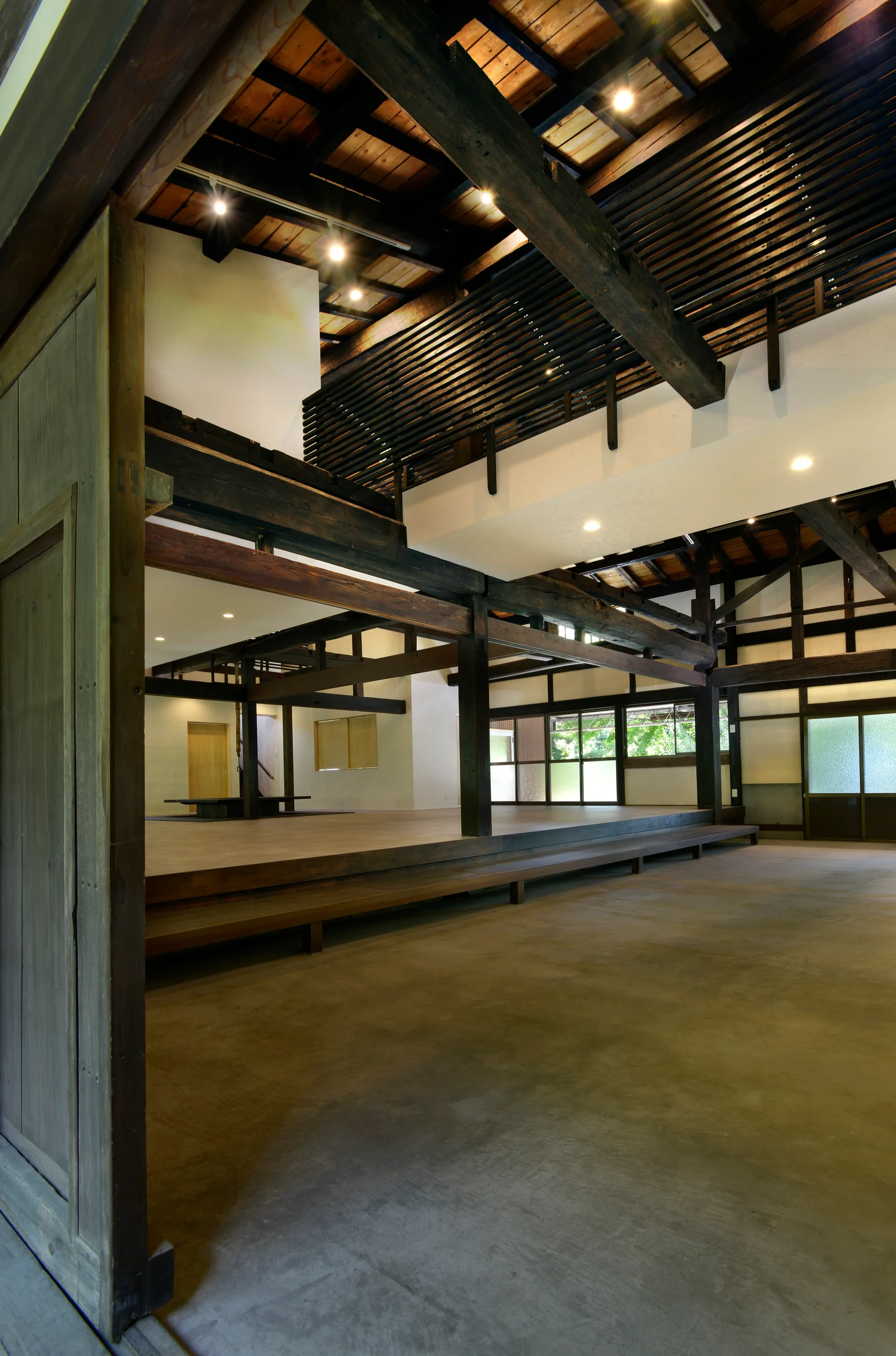 A spacious Japanese-style interior featuring a raised wooden platform floor, a grey concrete floor, and heavy dark-stained exposed timber ceiling beams.