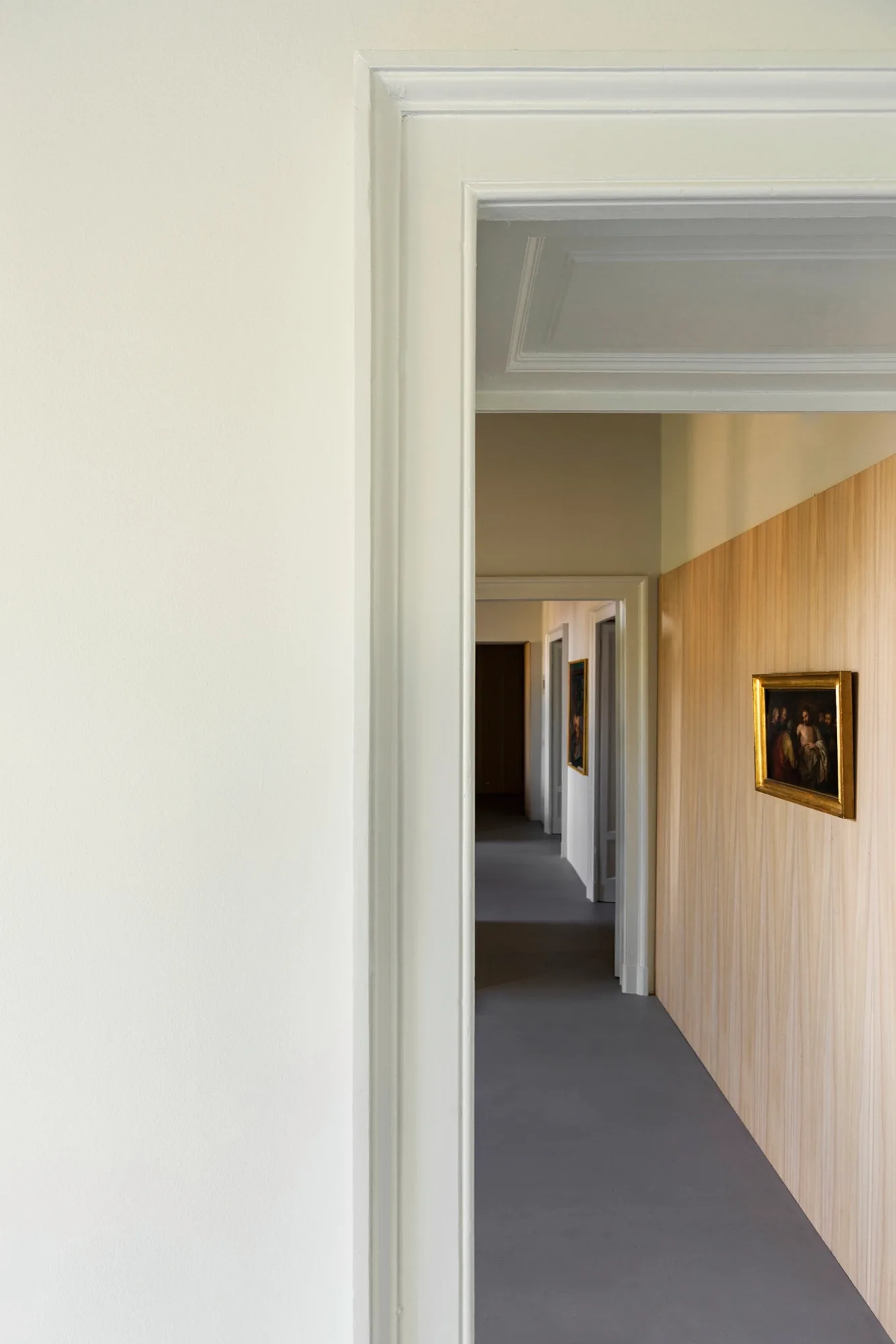 A view through white doorways down a long hallway featuring grey flooring, vertical wood wall paneling, and a small gold-framed painting.