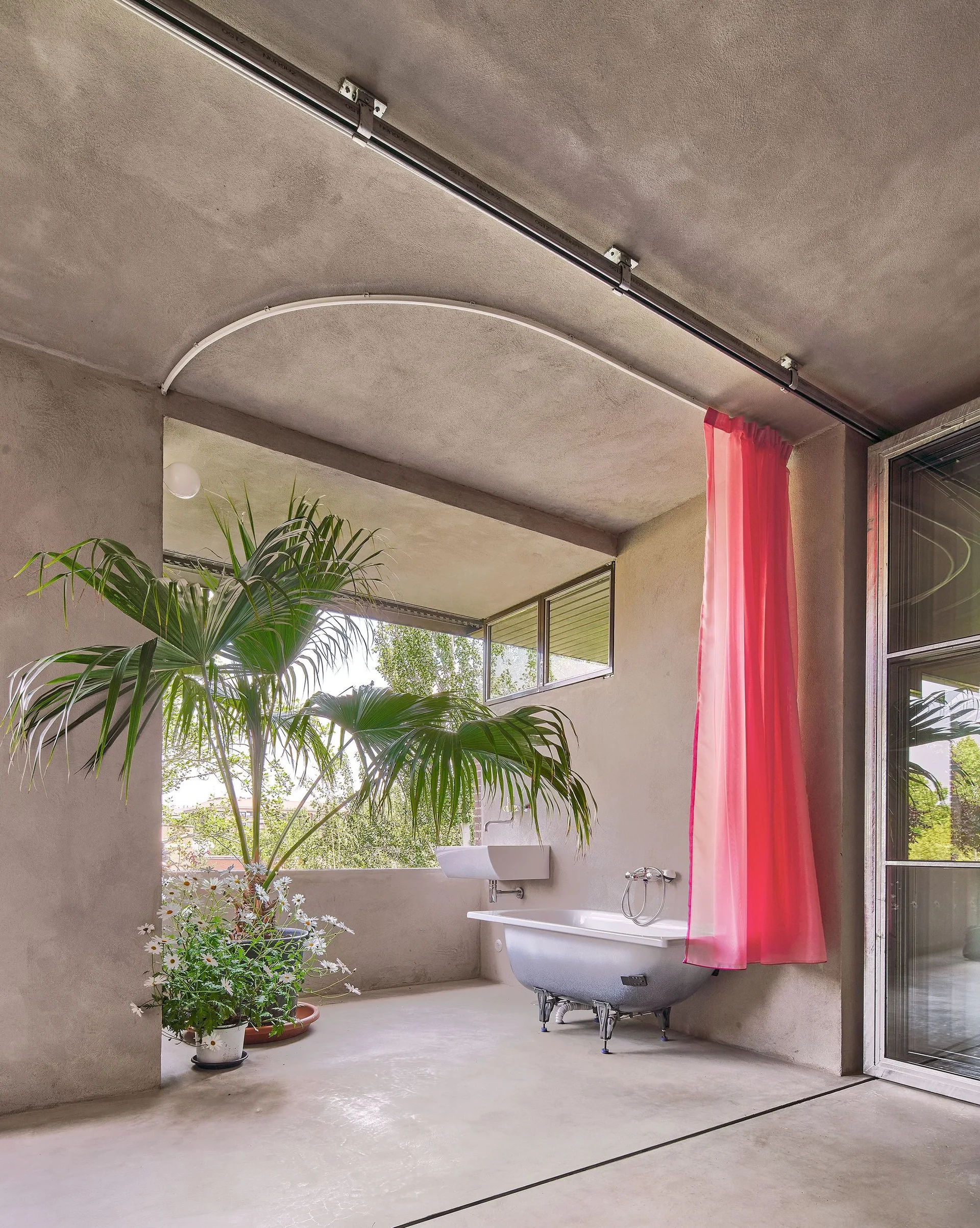A concrete bathroom features a freestanding bathtub, a wall-mounted sink, potted green plants, and a vibrant pink curtain hanging from a ceiling track.