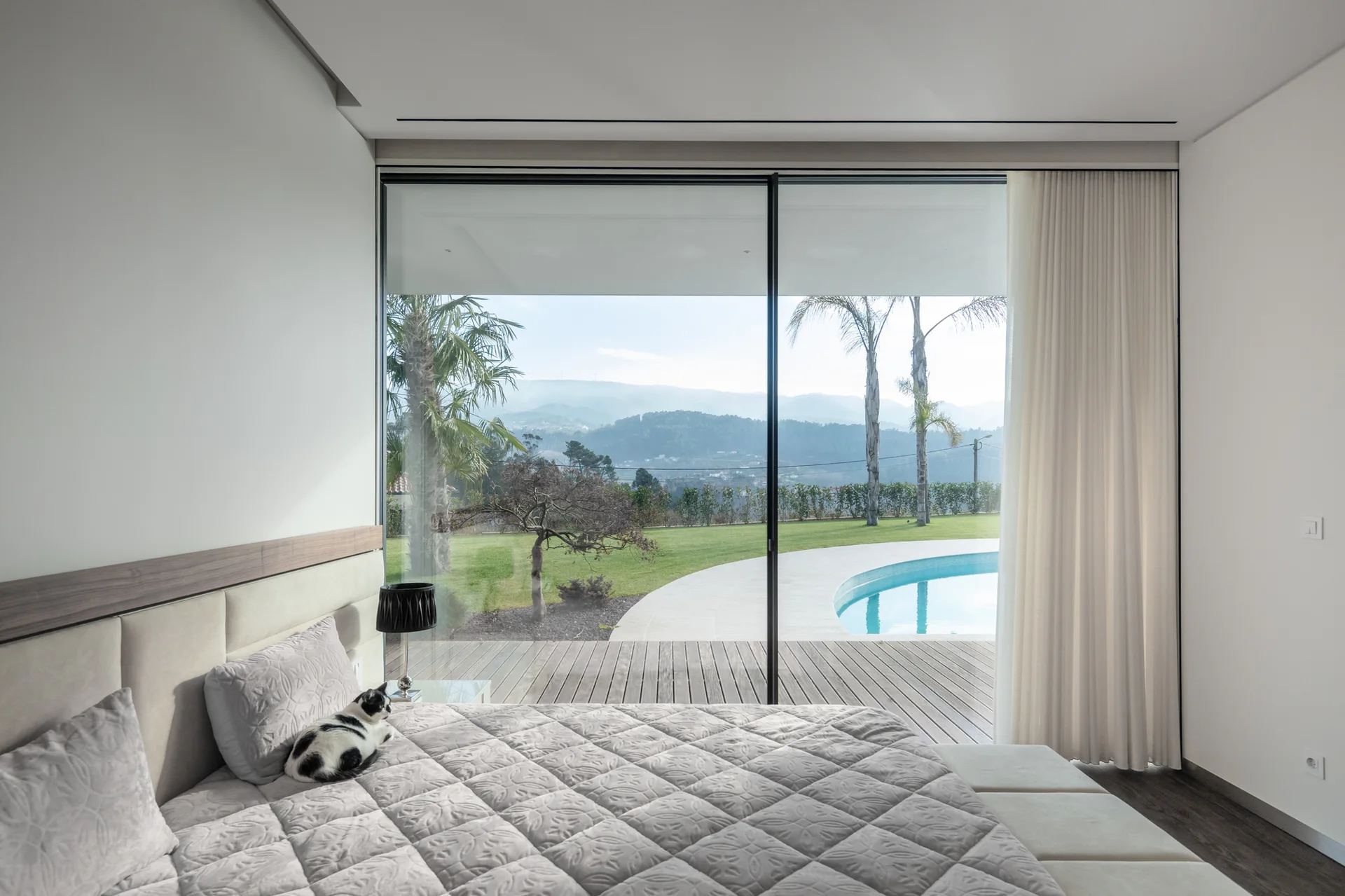 A minimalist bedroom featuring a grey quilted bed facing a large floor-to-ceiling sliding glass door that opens onto a wooden deck and backyard swimming pool.