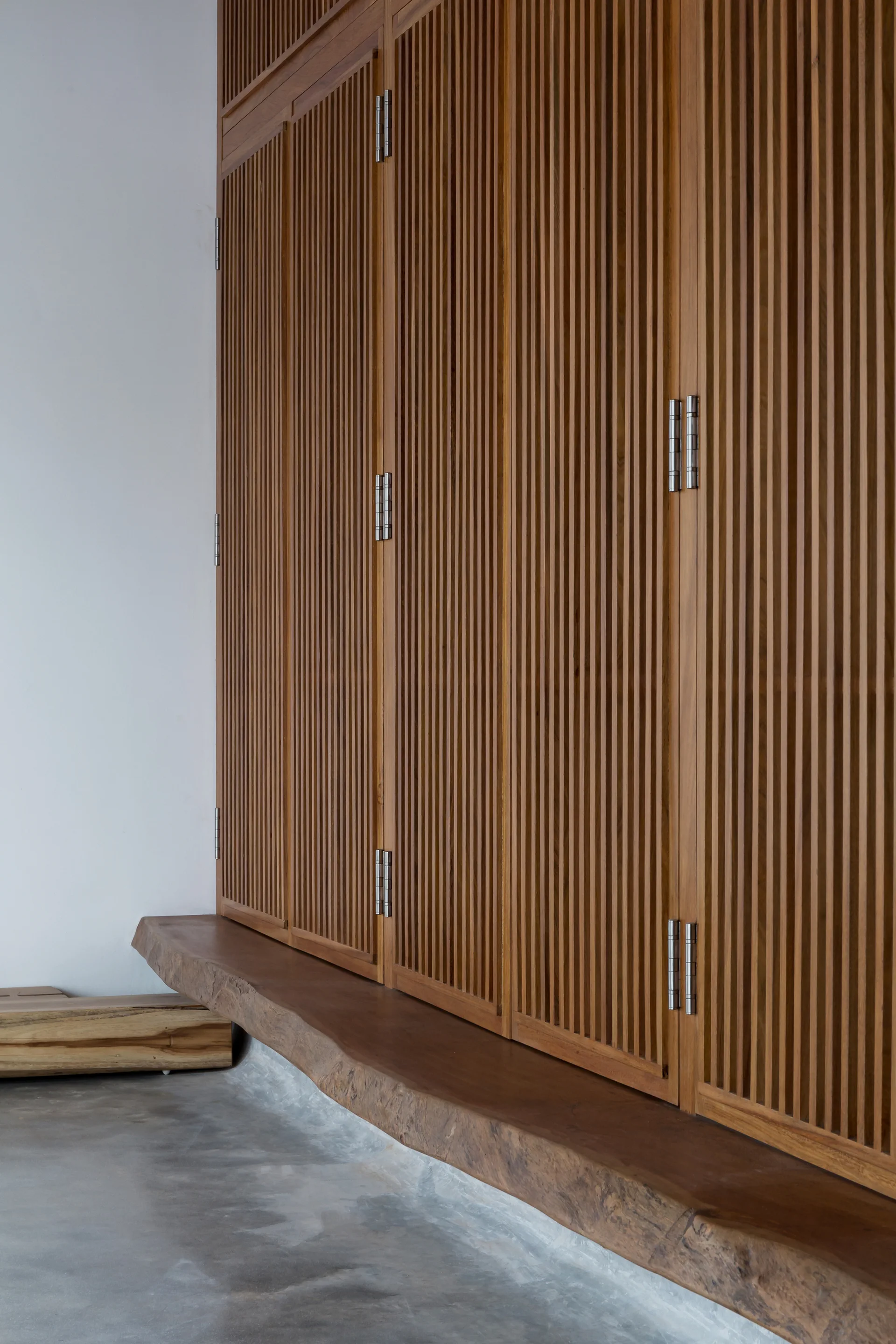 Full-height slatted wooden cabinets with silver hinges stand above a natural live-edge wood bench on a polished grey concrete floor.