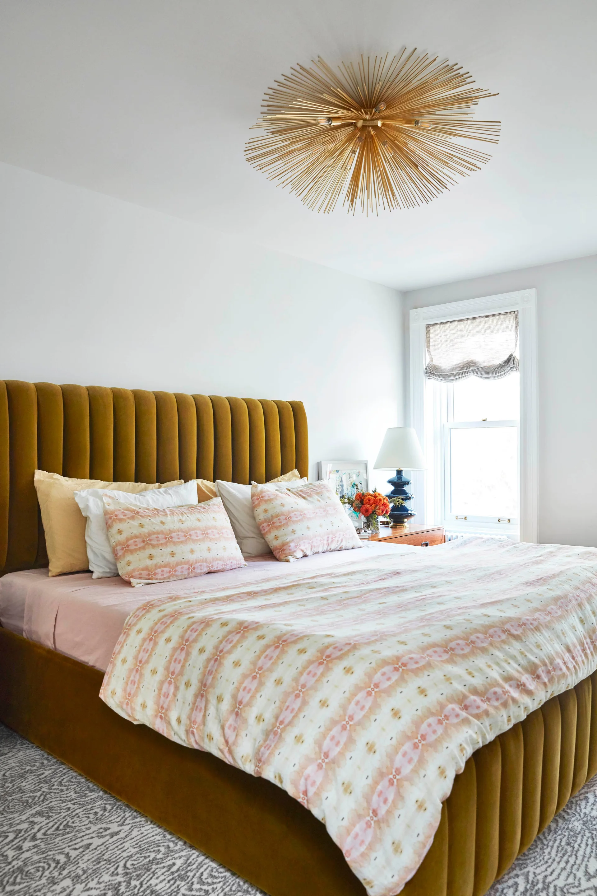 A bedroom features a mustard yellow velvet channel-tufted bed with ikat pattern bedding, a blue ceramic table lamp, and a gold sputnik ceiling fixture.