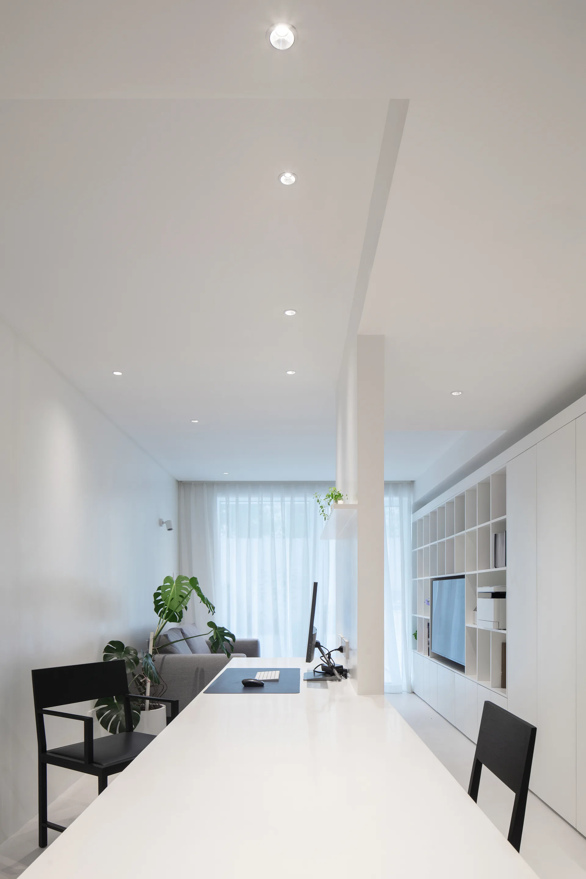 A minimalist white home office featuring a long peninsula desk, black chairs, a built-in shelving unit, and sheer curtains.