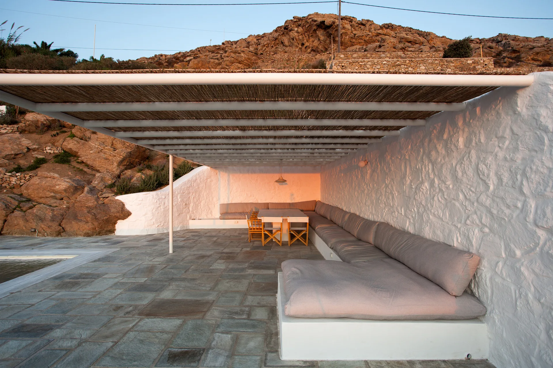 An outdoor patio features built-in white plaster bench seating with grey cushions under a thatched reed ceiling next to a stone tile floor.