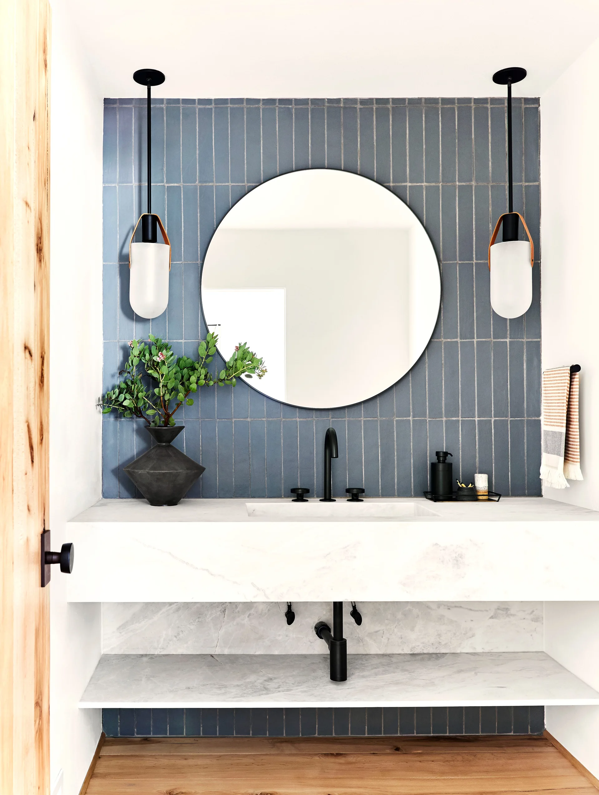 A modern bathroom vanity features a floating marble countertop, a round mirror, and two hanging lights against a blue vertical tile backsplash.
