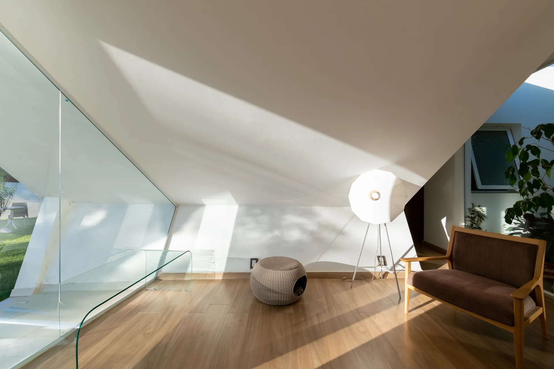 A minimalist attic living room features a slanted white ceiling, a brown armchair, a knit ottoman, and a glass table by a large window.