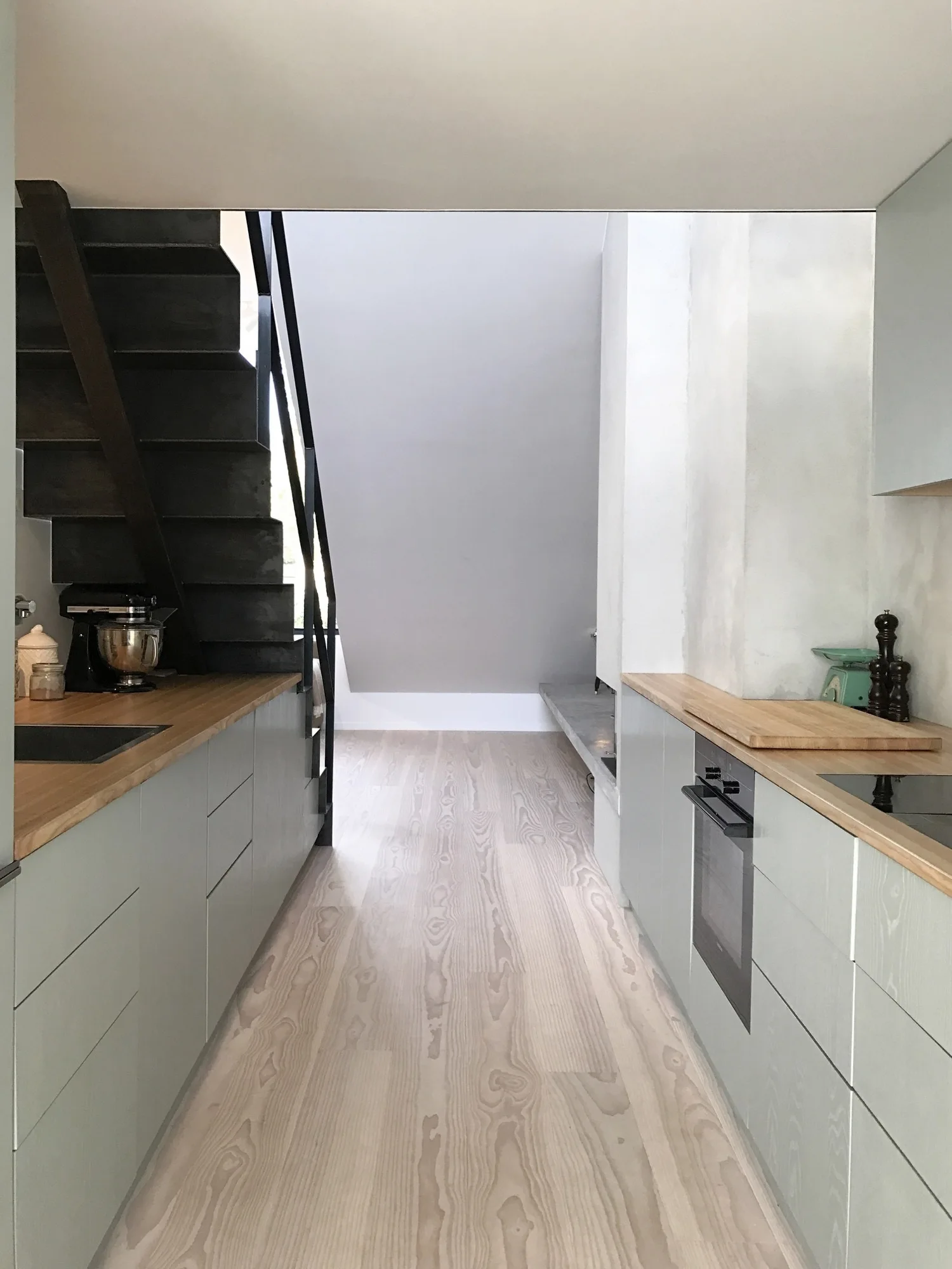 A narrow galley kitchen features sage green slab-front cabinets, light wood countertops, wood flooring, and a dark metal staircase on the left.