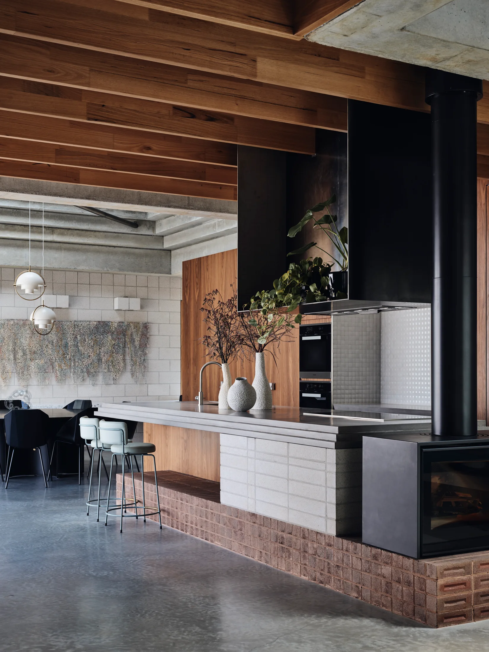 An industrial kitchen features a concrete-topped island with a red brick base, built-in fireplace, and exposed wood ceiling beams.