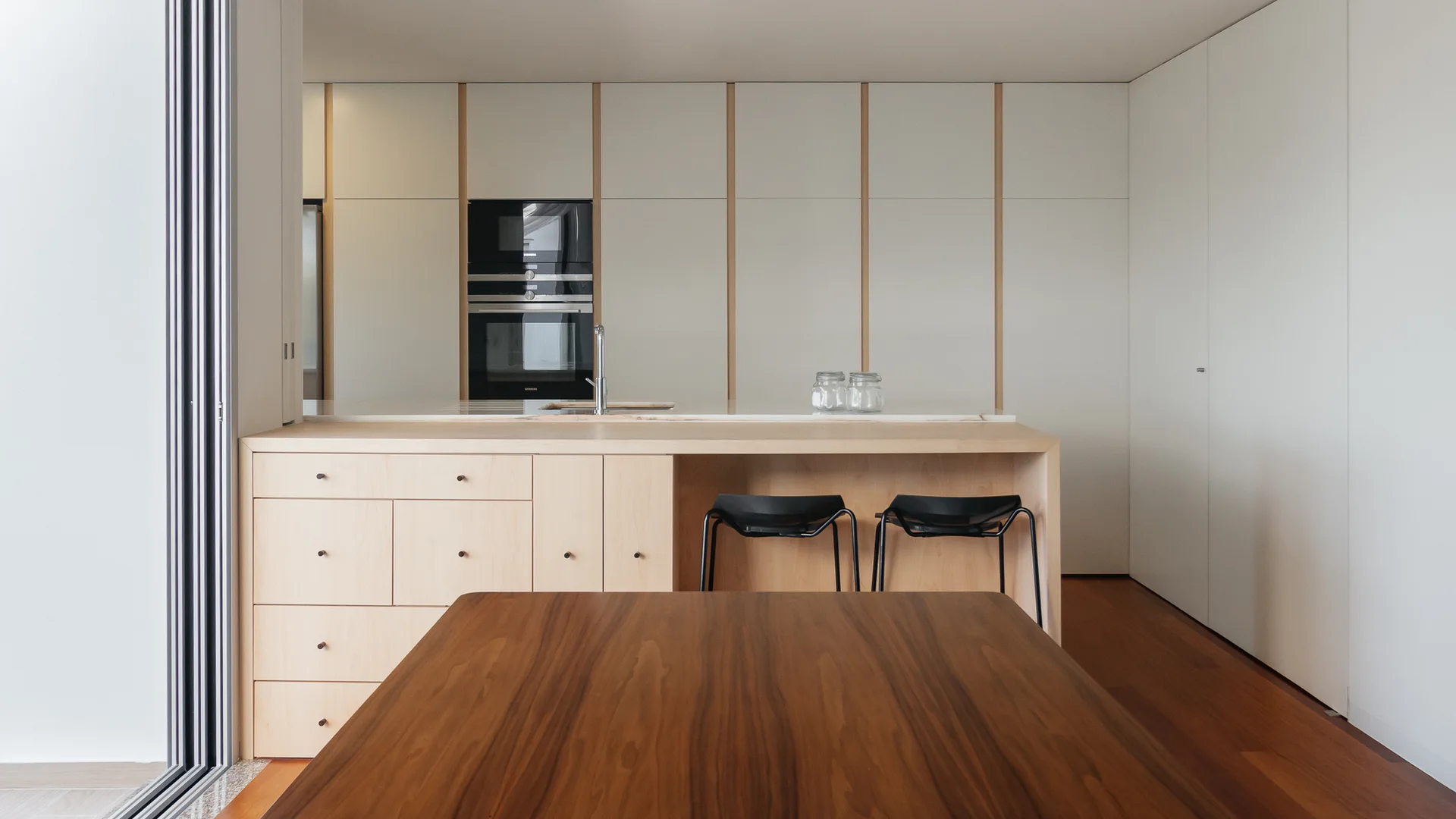 A modern kitchen features a light wood island with black stools, white floor-to-ceiling cabinets, and a dark wood table in the foreground.