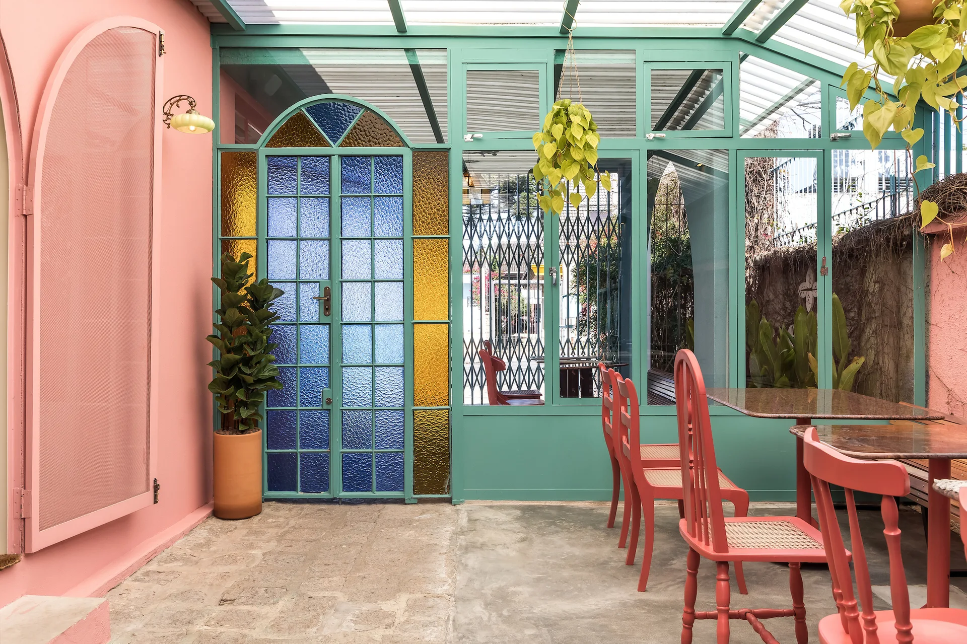 A covered outdoor patio featuring salmon pink walls, a teal-framed glass roof, a blue and gold stained-glass door, and red dining chairs.