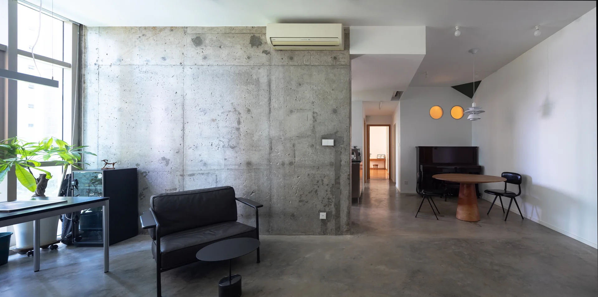 An industrial apartment interior featuring a raw concrete wall, polished concrete floors, a dark leather sofa, and a round wooden dining table.