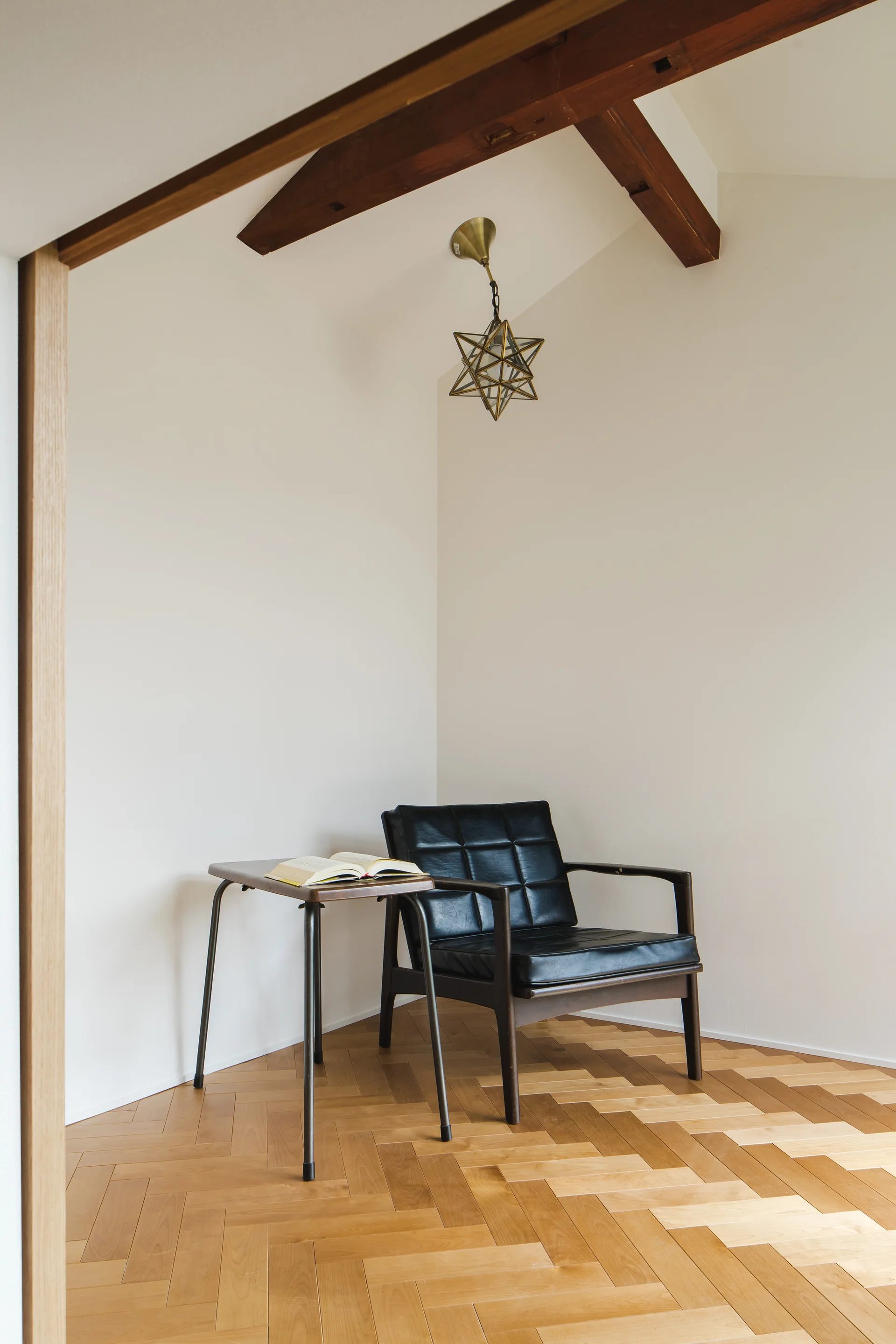 A black tufted leather armchair and a small wood side table sit on a herringbone wood floor beneath an exposed ceiling beam and a star pendant.
