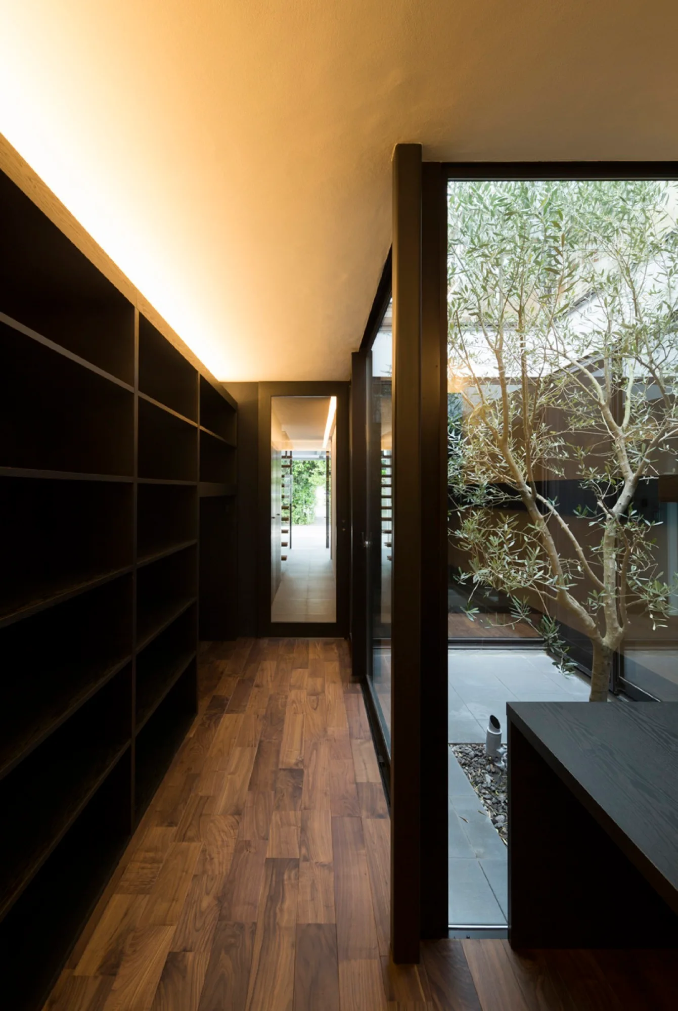 A minimalist hallway features black open shelving, wood floors, and floor-to-ceiling glass looking out onto a central courtyard with a small tree.