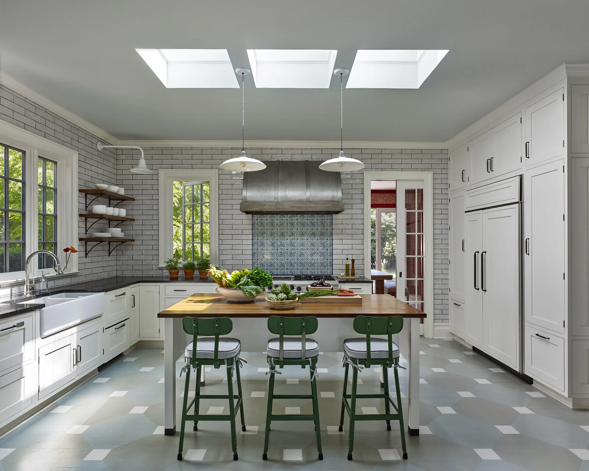 A bright kitchen features a wooden island with green stools under three skylights, surrounded by white cabinets and gray tiled walls.
