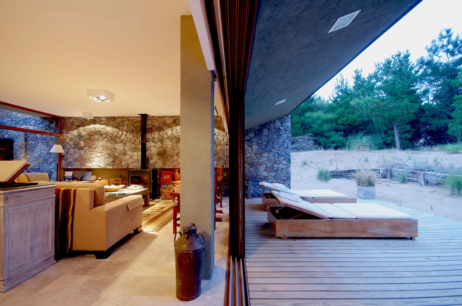 An indoor living area with a stone wall and fireplace transitions through a sliding door to a wooden deck with wooden chaise lounges.