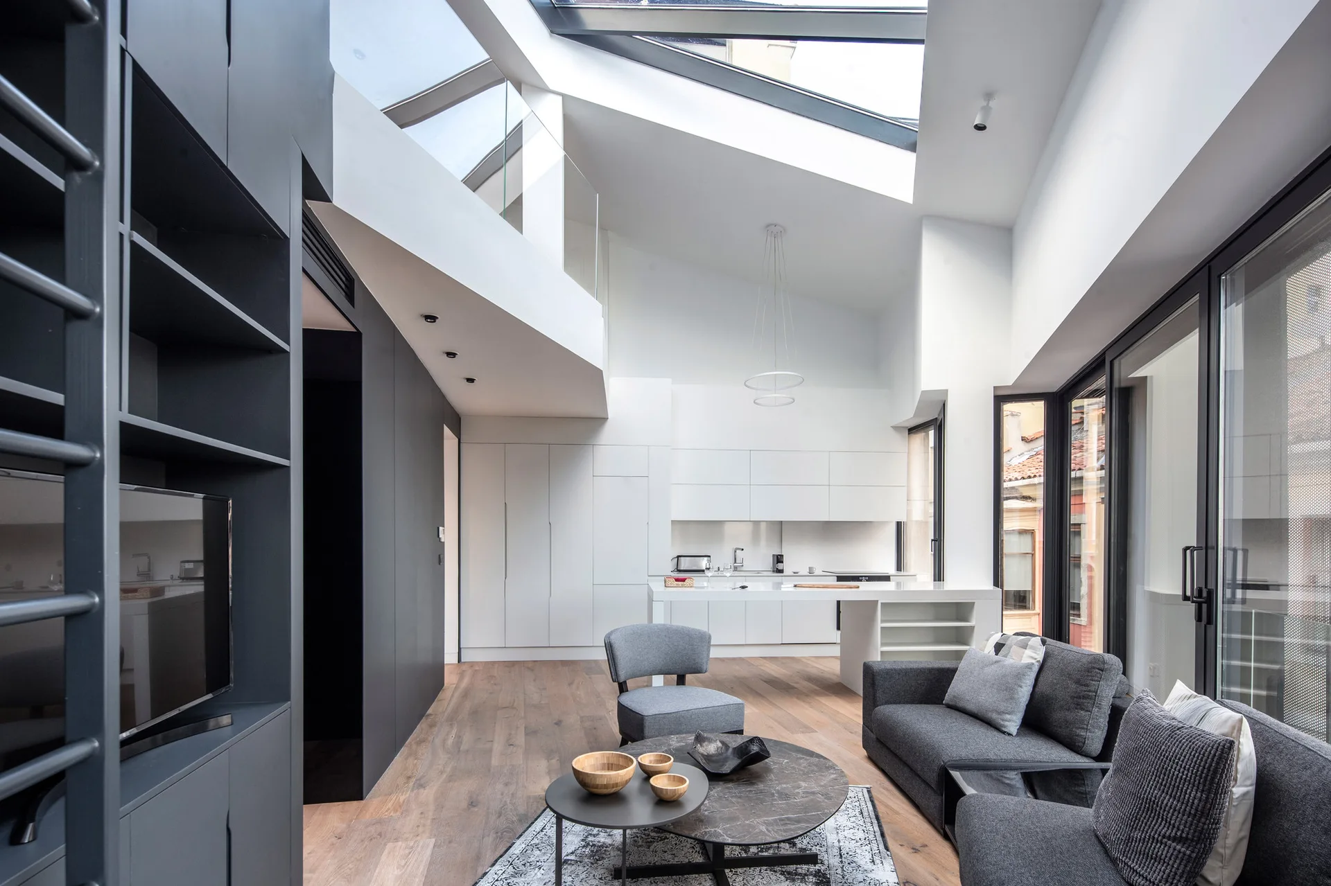 Modern open-plan living room featuring a grey sofa, marble coffee table, high ceiling with large slanted skylights, and a dark grey storage wall.