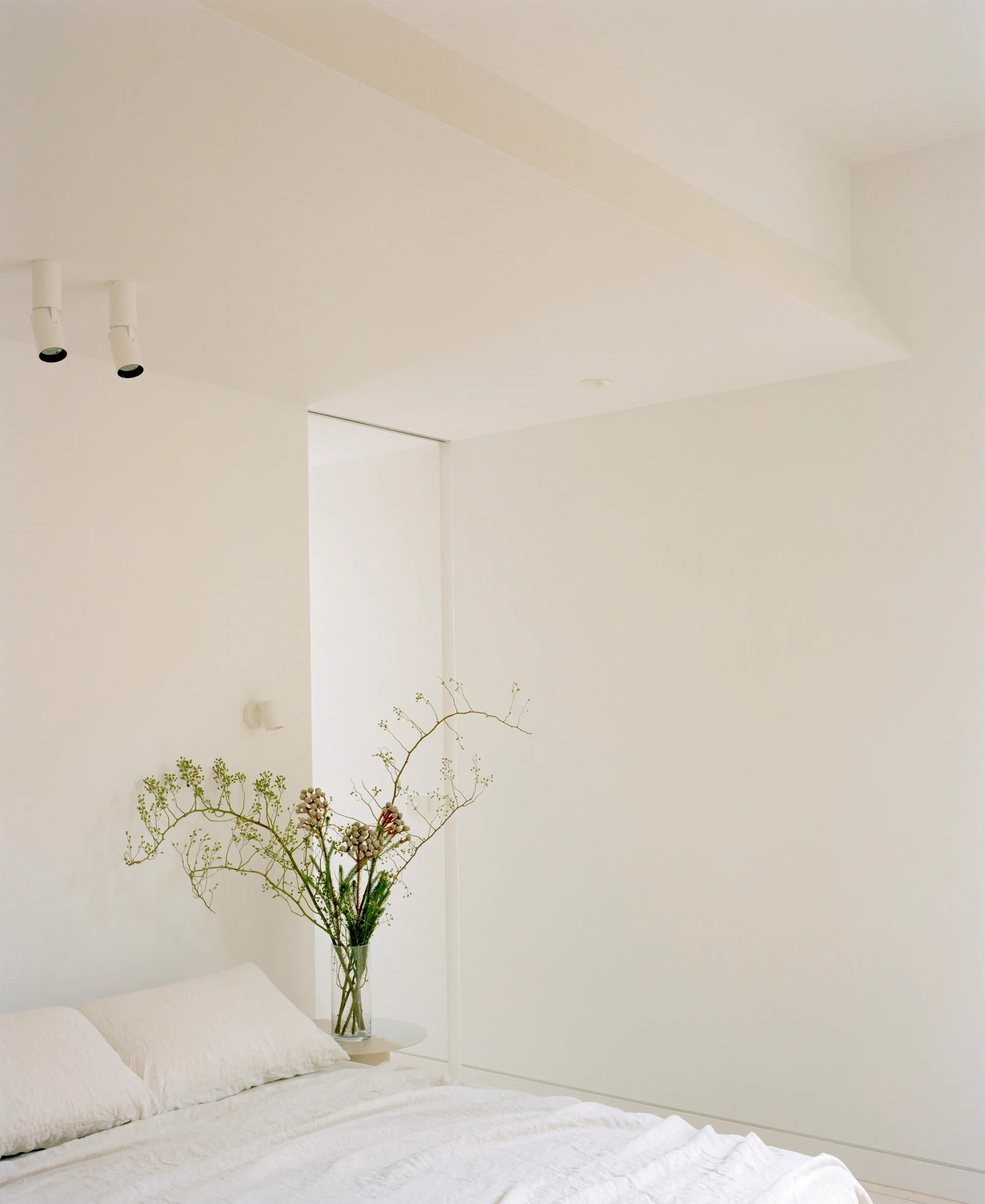 A minimalist white bedroom features white bed linens, a small side table holding a tall glass vase of branches, and spotlights on the ceiling.