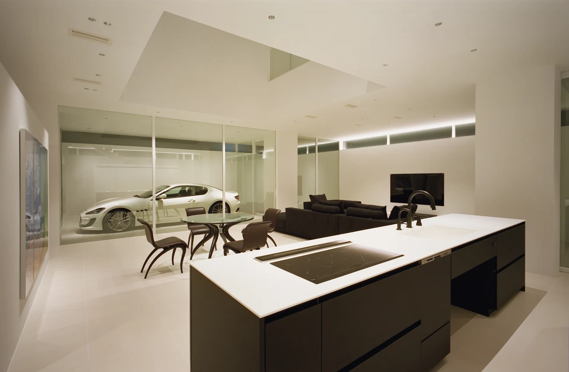 A minimalist open-plan kitchen and living room featuring a black kitchen island, a dark dining set, and a sports car visible behind a glass wall.