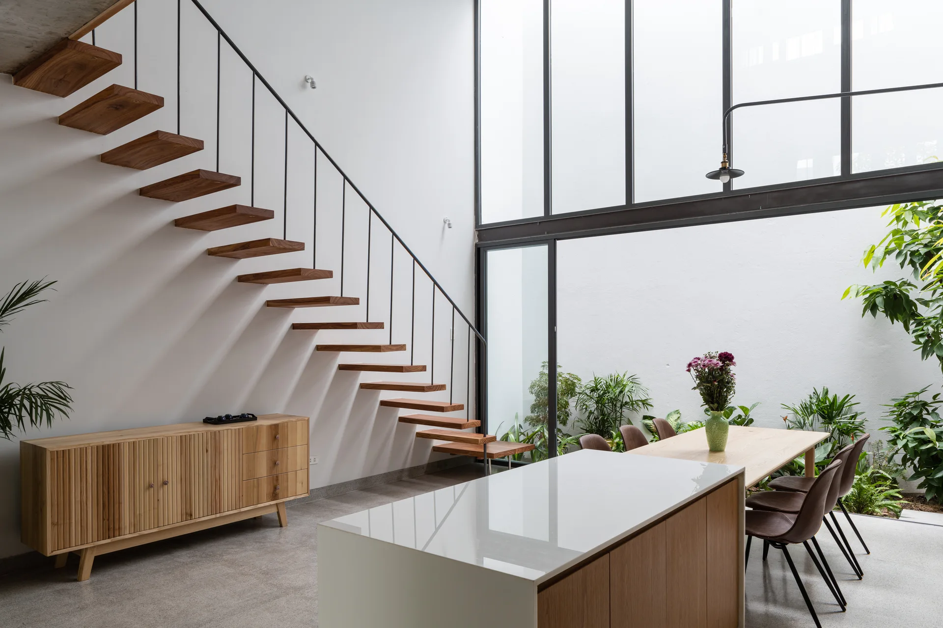 A modern dining area features a floating wooden staircase, a slatted sideboard, and a large glass window wall looking out to a small indoor garden.