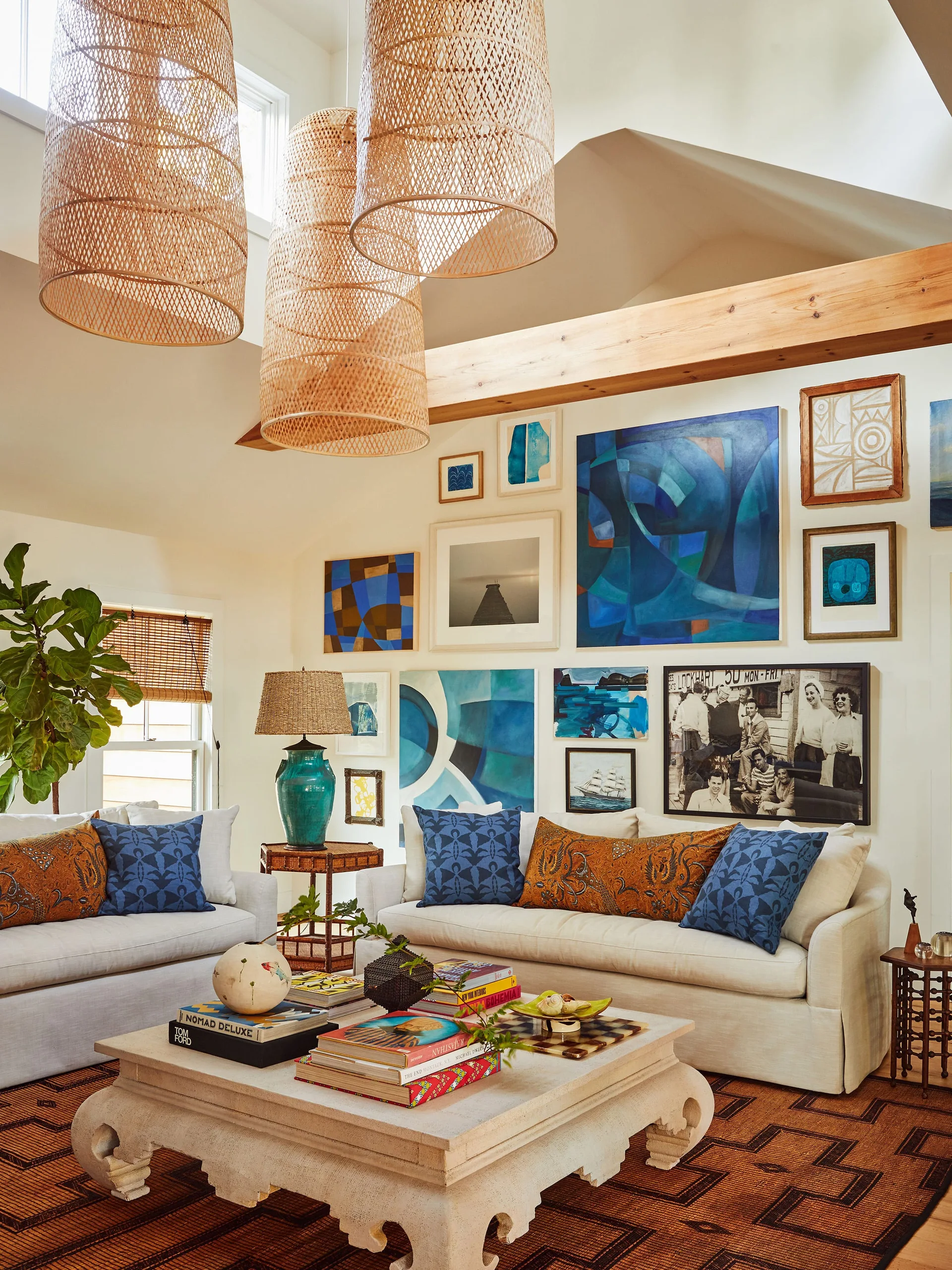 A bright living room features two cream sofas, a large gallery wall of blue art, a carved wood coffee table, and three oversized woven pendant lights.