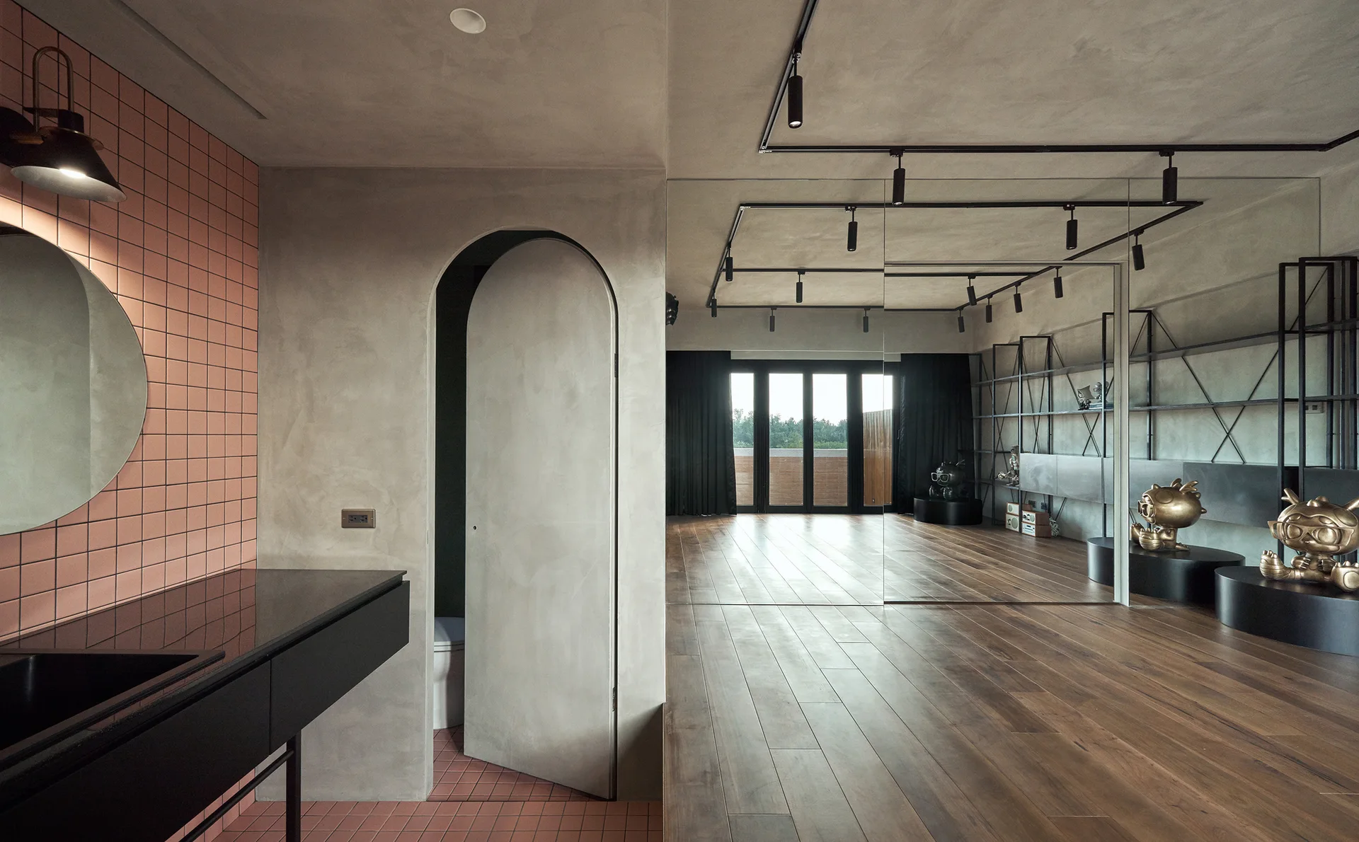 A modern bathroom features pink square tiles, a circular mirror, a concrete-textured arched door, and wood flooring in the adjacent room.