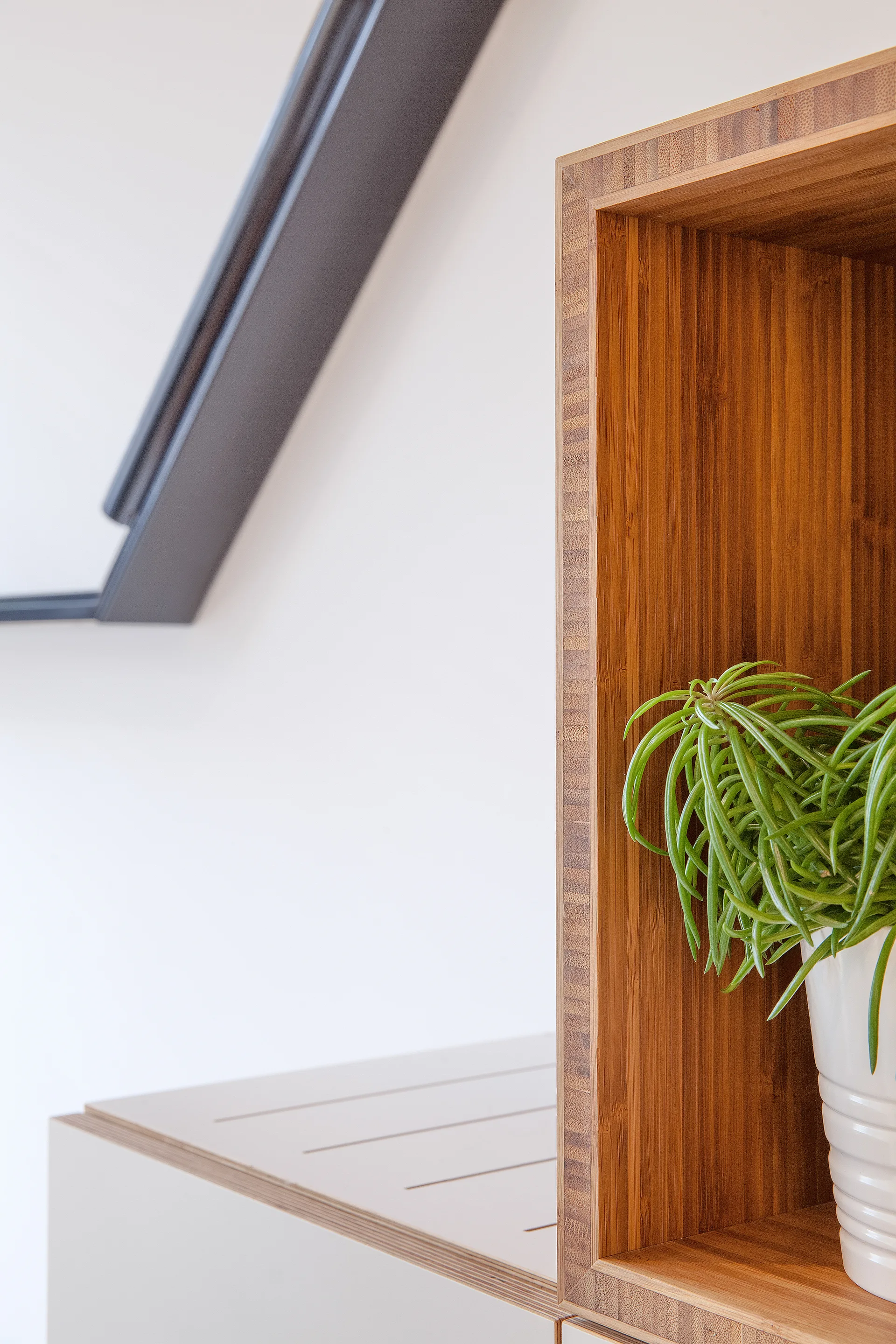 A close-up of a warm wood bamboo shelving unit holding a white ribbed pot with a green plant next to an angled skylight window.