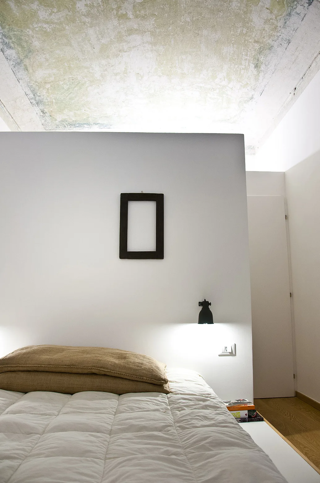 A minimalist bedroom features a quilted white bed, textured hessian pillows, a black wall lamp, and a distressed vaulted ceiling.