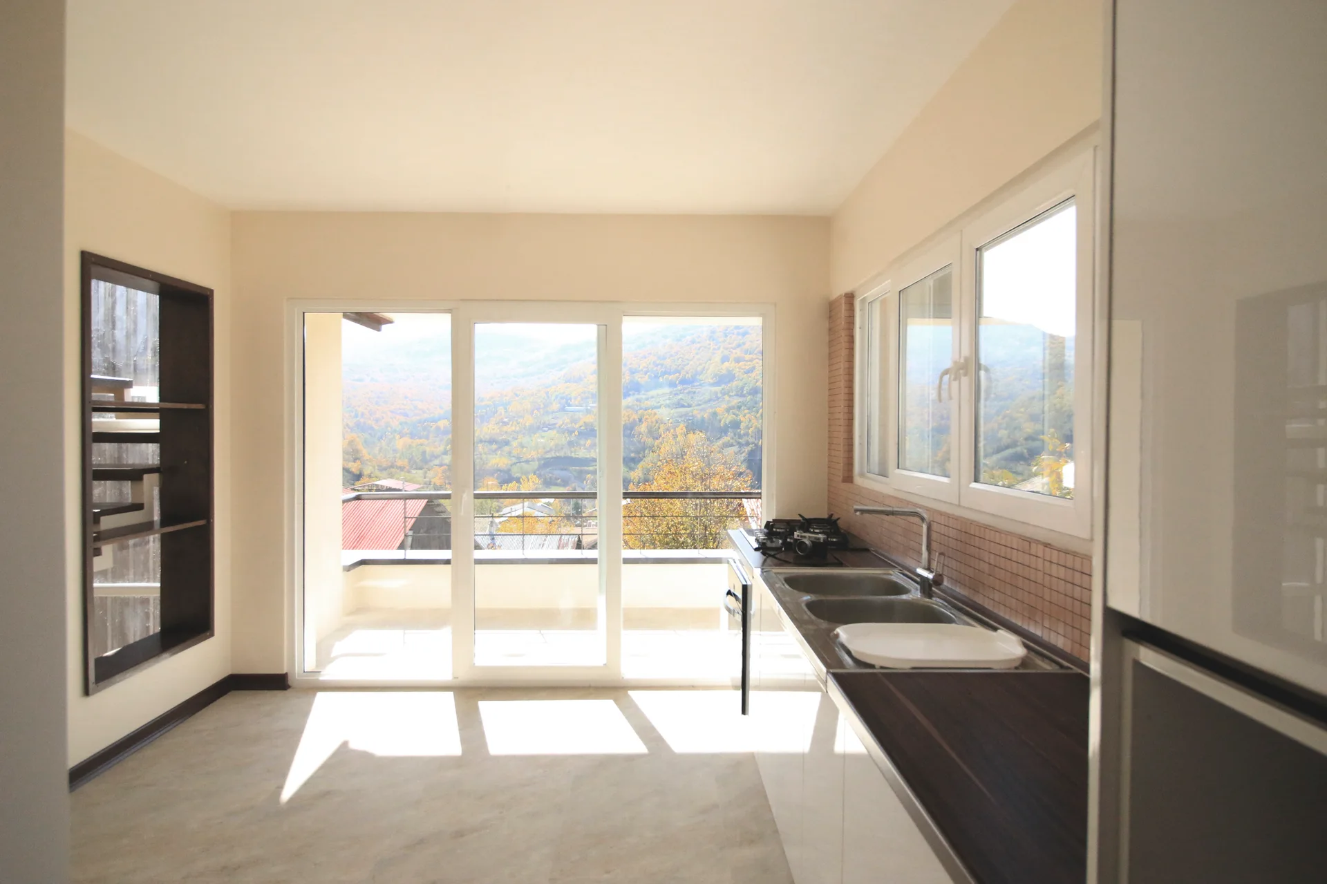 A bright minimalist kitchen features glossy white cabinets, a dark wood-toned countertop, a tile backsplash, and sliding glass doors to a balcony.