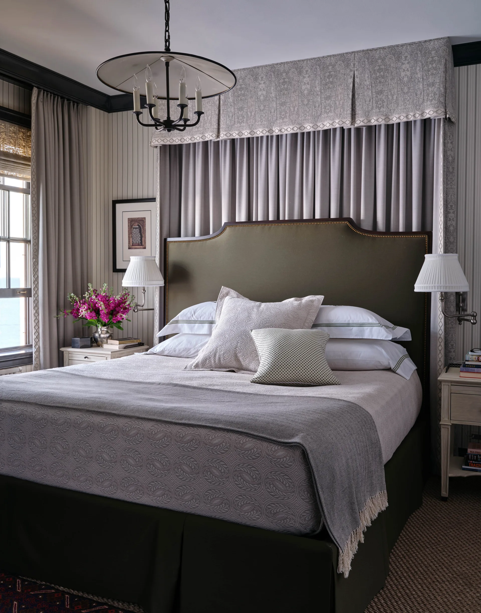 A traditional bedroom features a bed with an olive green headboard, a patterned valance, and bedside tables with sconces.