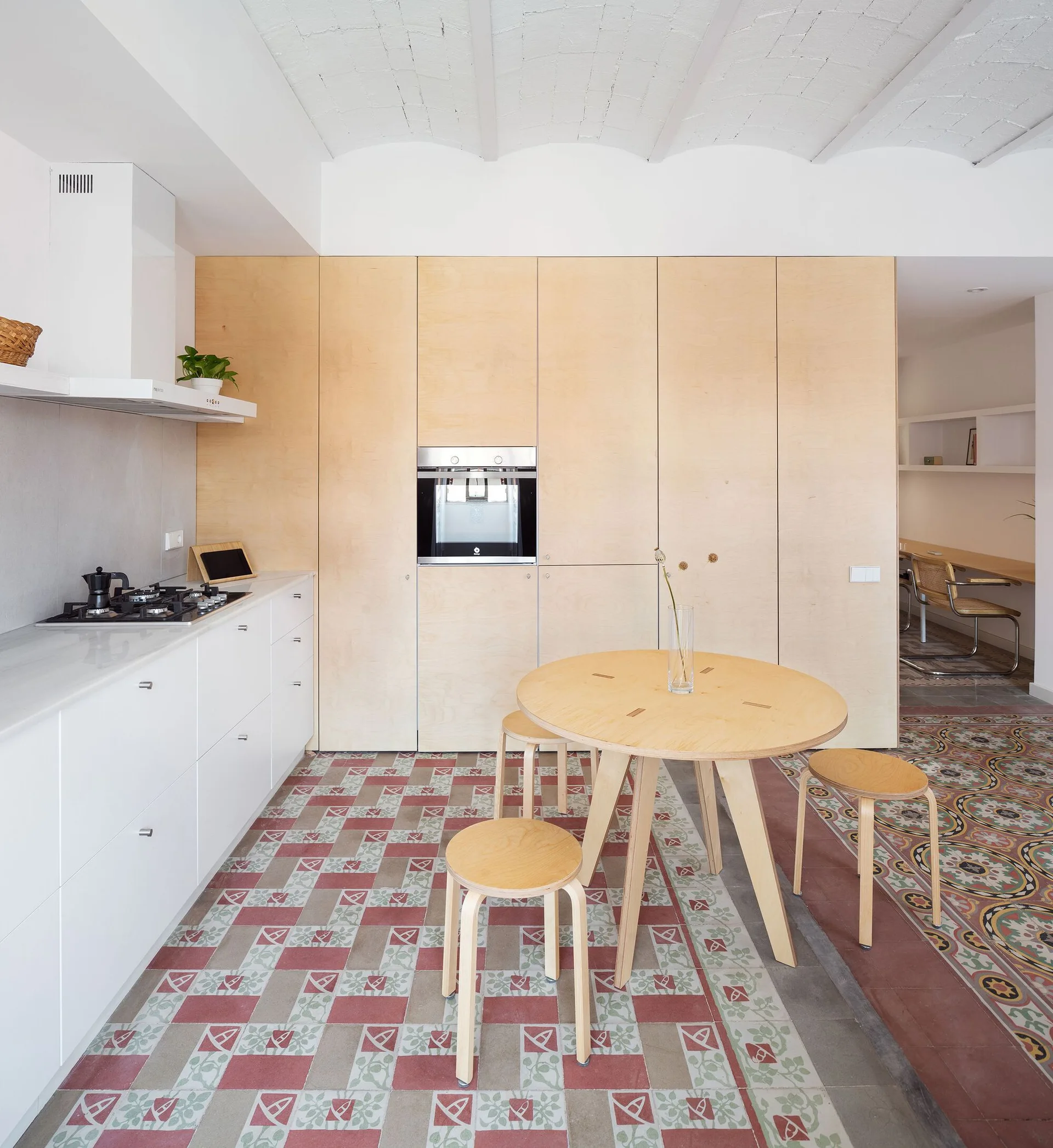 A modern kitchen with full-height plywood cabinets, a round wooden dining table with matching stools, and patterned red and green floor tiles.