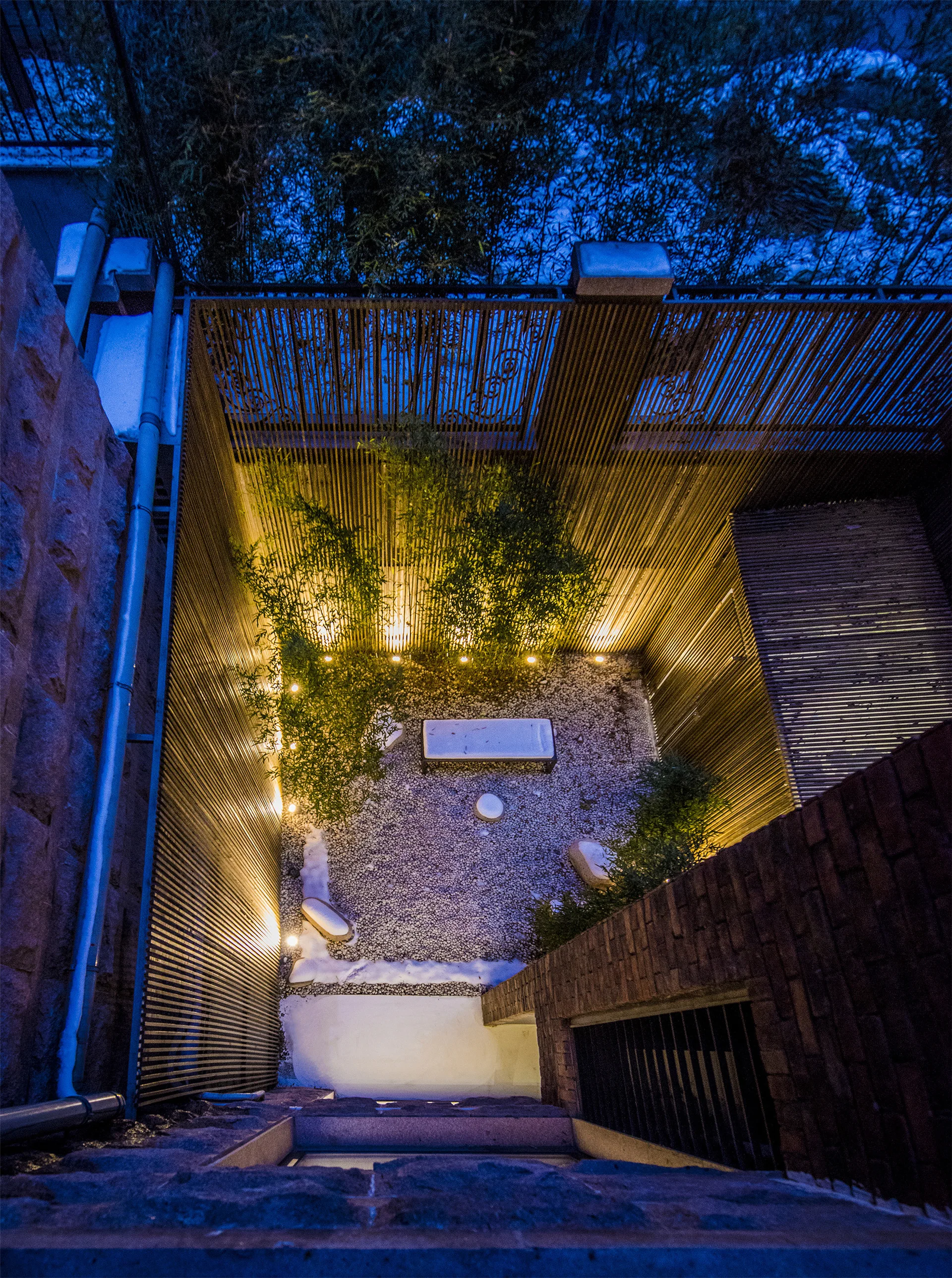 An overhead night view of a small enclosed courtyard with vertical wood slat walls, gravel ground, and warm ground lighting highlighting a central bench.