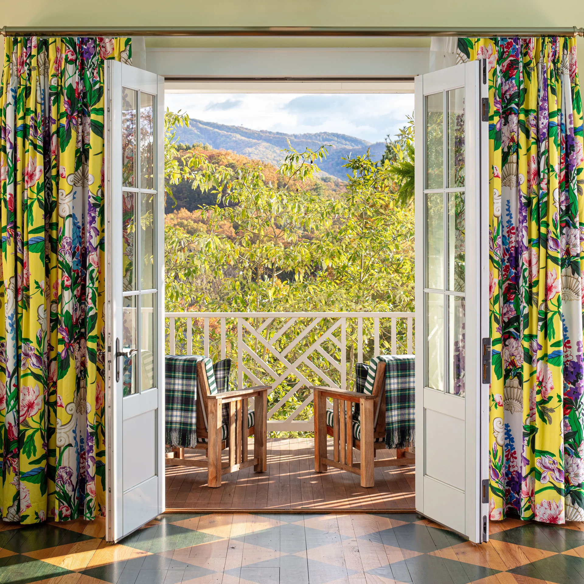 A view through open white French doors to a wooden balcony with two chairs overlooking a green mountain valley, framed by yellow floral curtains.