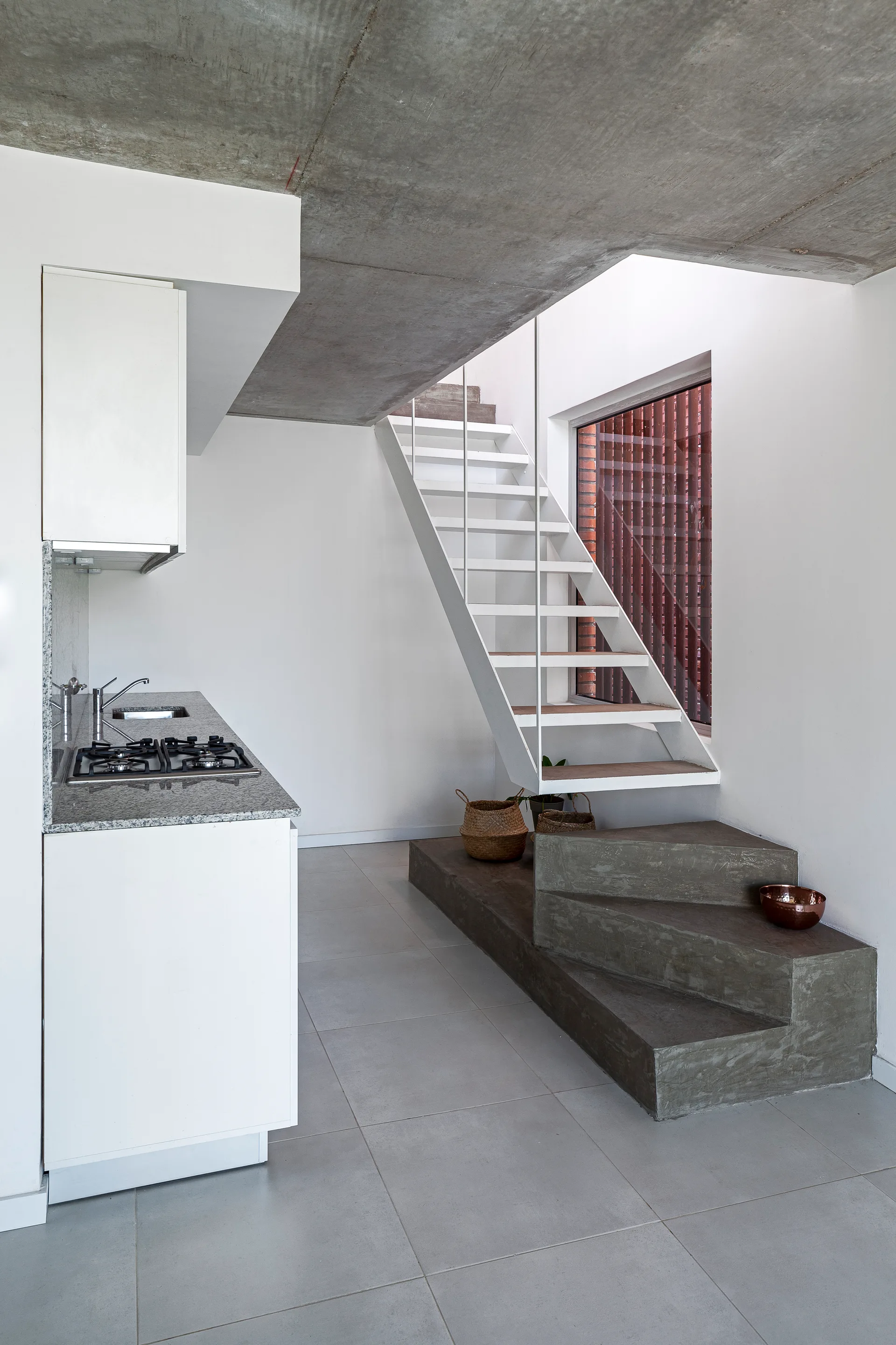 A minimalist white metal staircase rises from a solid concrete base next to a white kitchen counter under a concrete ceiling.