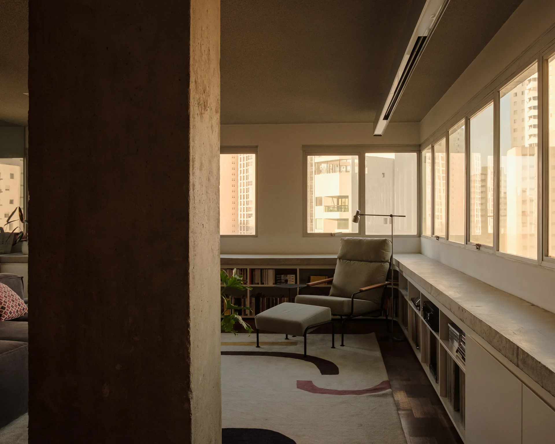 A modern lounge area featuring a concrete column in the foreground, a grey armchair with an ottoman, and low built-in shelving beneath a row of windows.