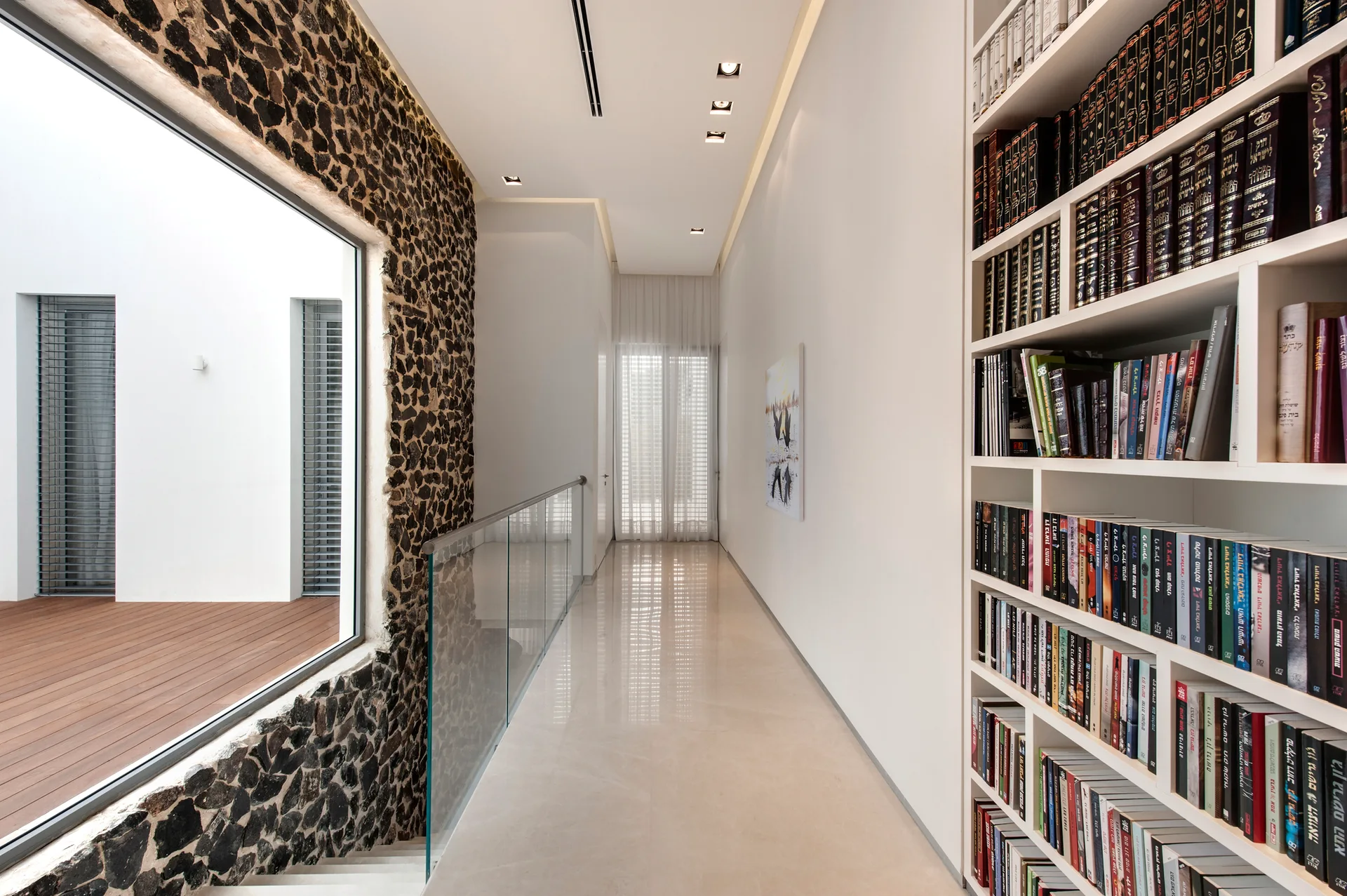 A bright hallway featuring a textured stone wall with a large window, a glass stair railing, and a tall white built-in bookshelf filled with books.