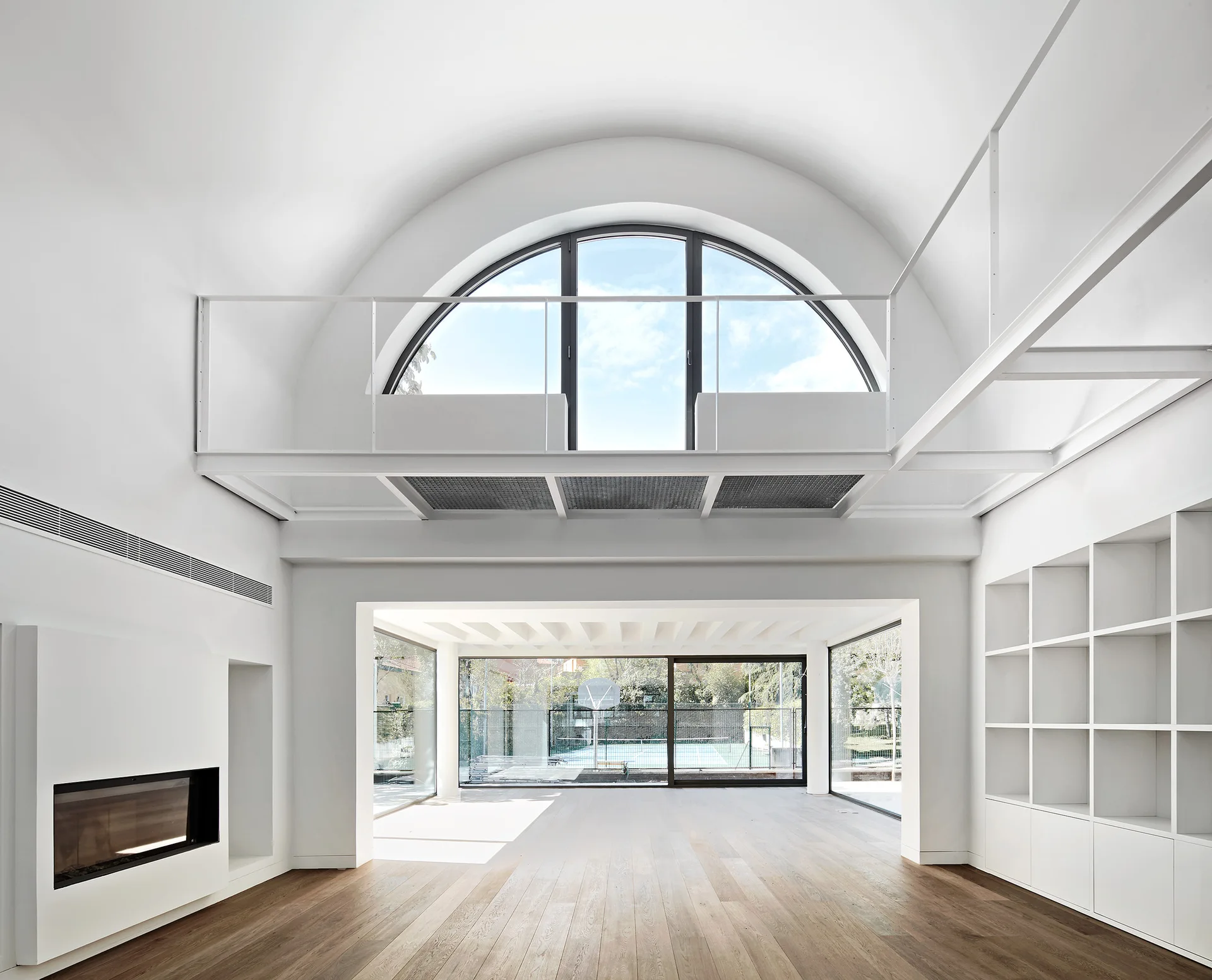 An empty minimalist living room with a double-height white ceiling, a large semi-circular arched window above a mezzanine, and a built-in bookshelf.