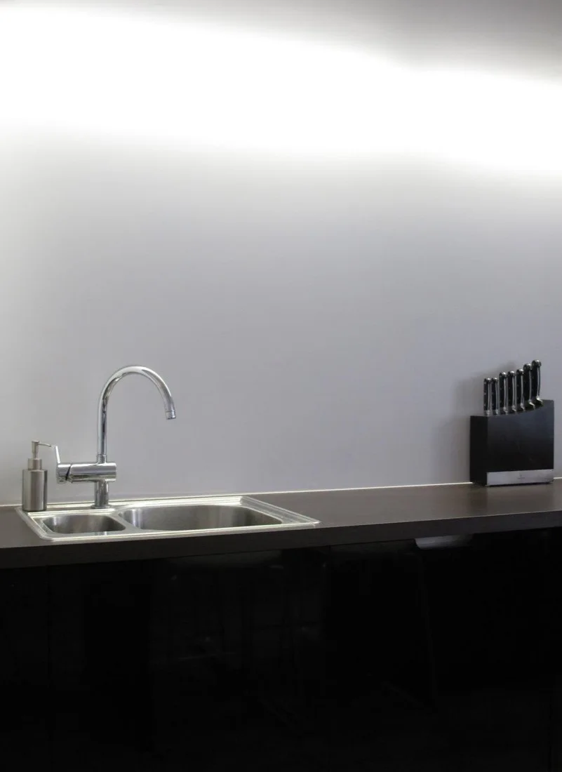 A stainless steel kitchen sink with a curved chrome faucet sits on a dark brown countertop against a smooth, light gray wall.