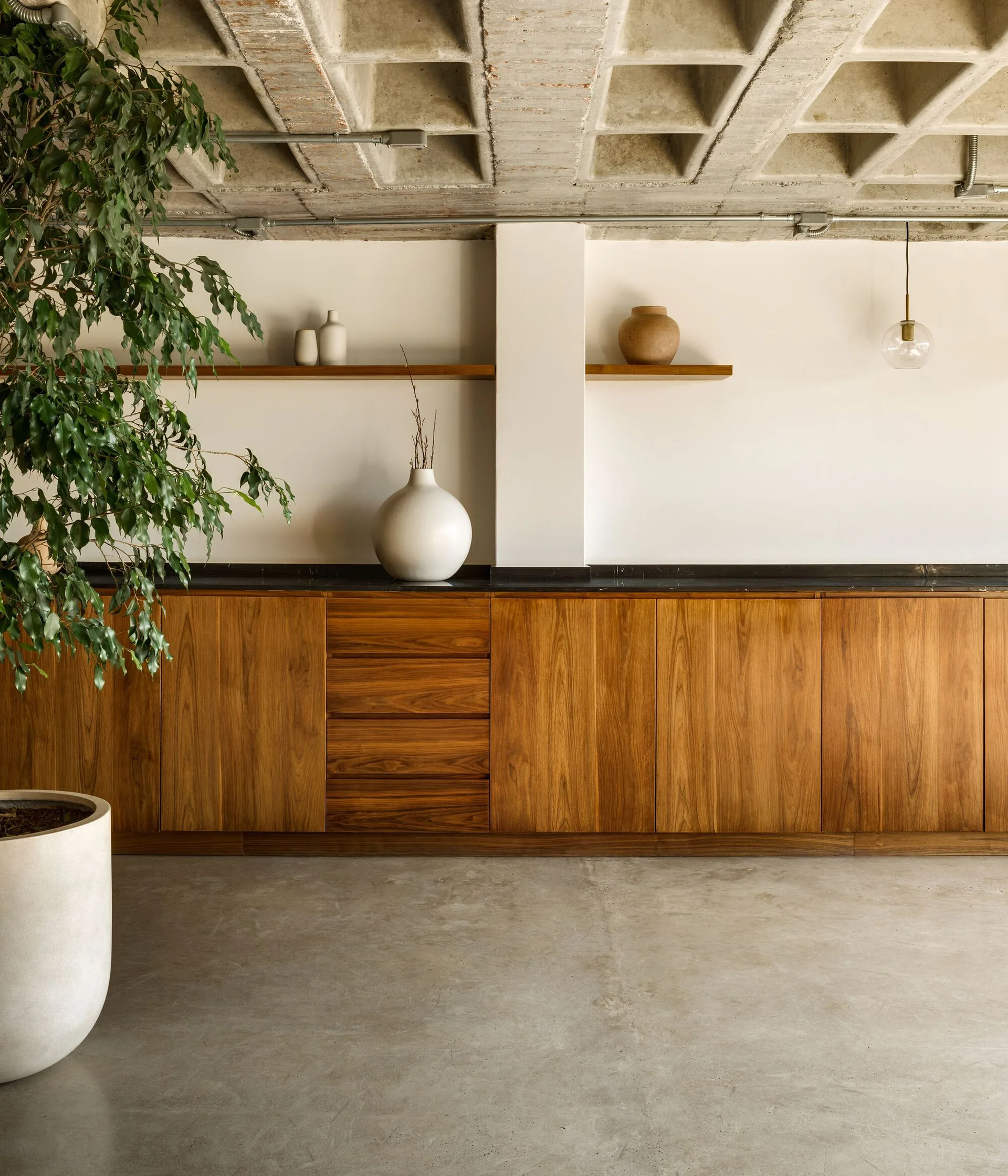A long wooden storage cabinet sits beneath an exposed concrete waffle slab ceiling and simple floating shelves displaying ceramic vases.
