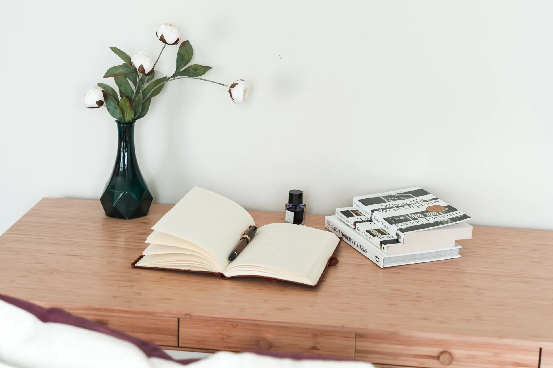 A light wood desk featuring an open lined notebook with a fountain pen, a small stack of books, and a dark green glass vase.