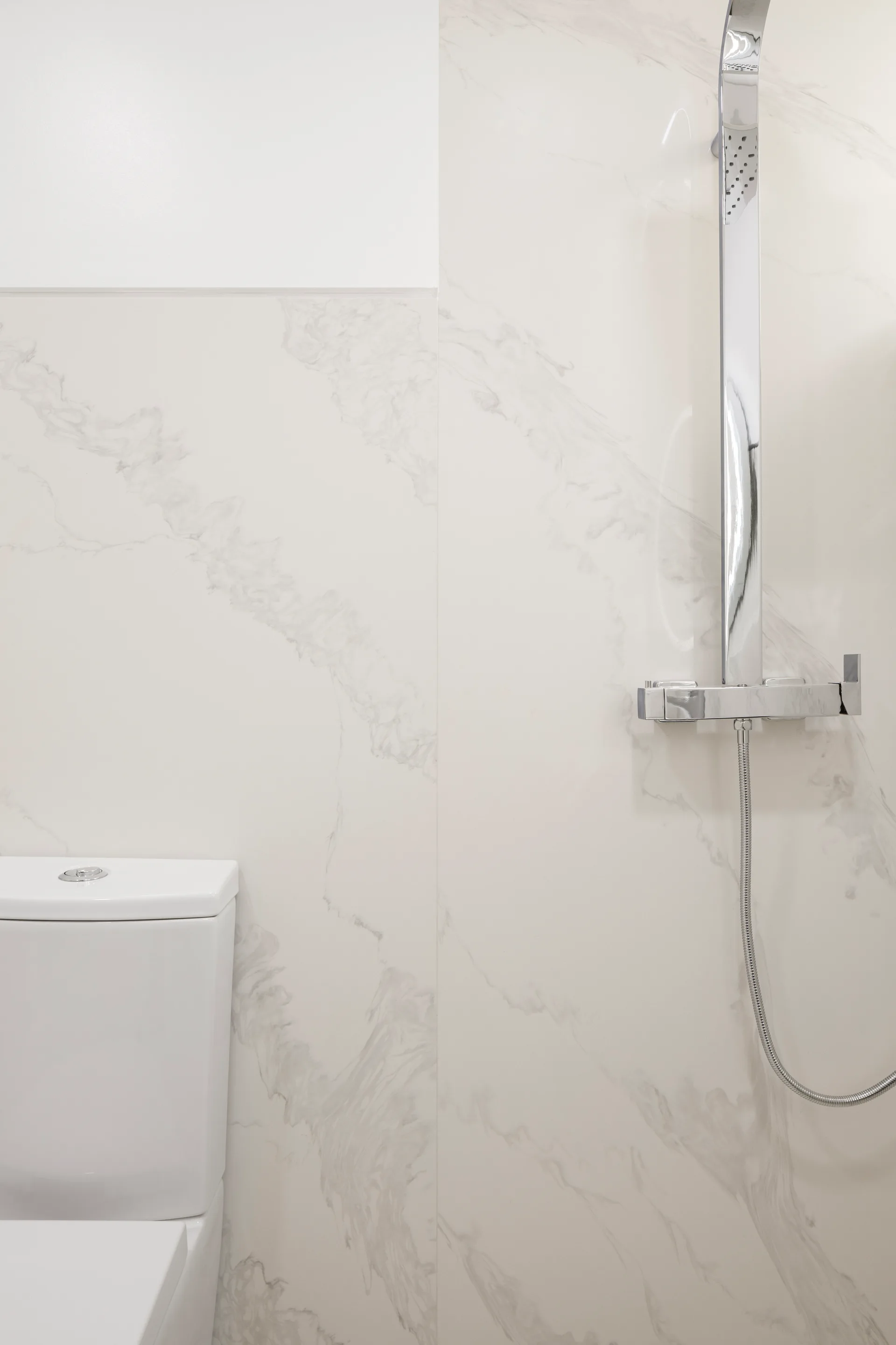 A bathroom wall tiled in large-format white marble with grey veining, featuring a chrome shower column and a white ceramic toilet tank.