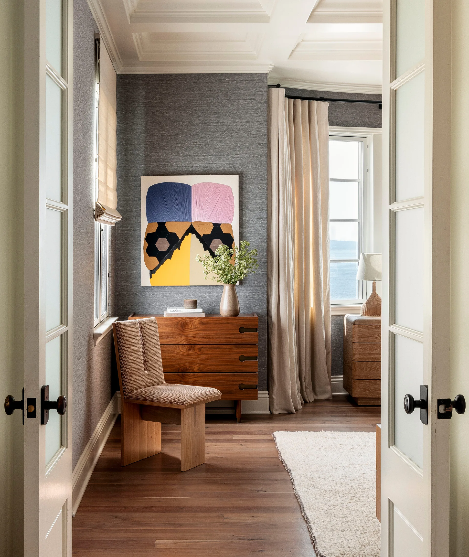 A bedroom corner viewed through white double doors, featuring charcoal textured wallpaper, a wooden dresser, a structured chair, and an abstract painting.