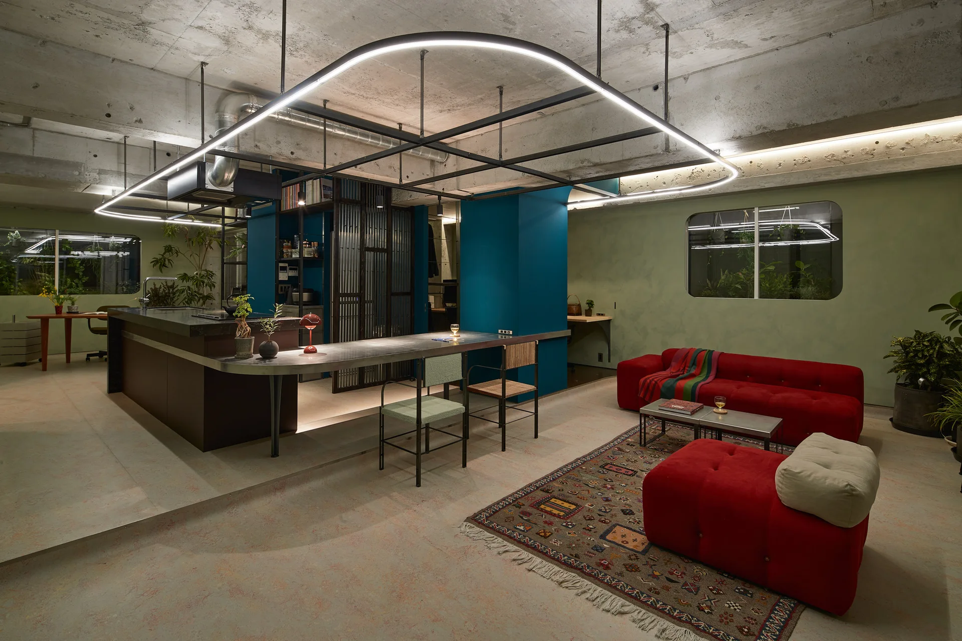 An open-plan loft features a bright red sofa, a patterned area rug, a dark kitchen island, and an overhead rectangular light frame under a concrete ceiling.