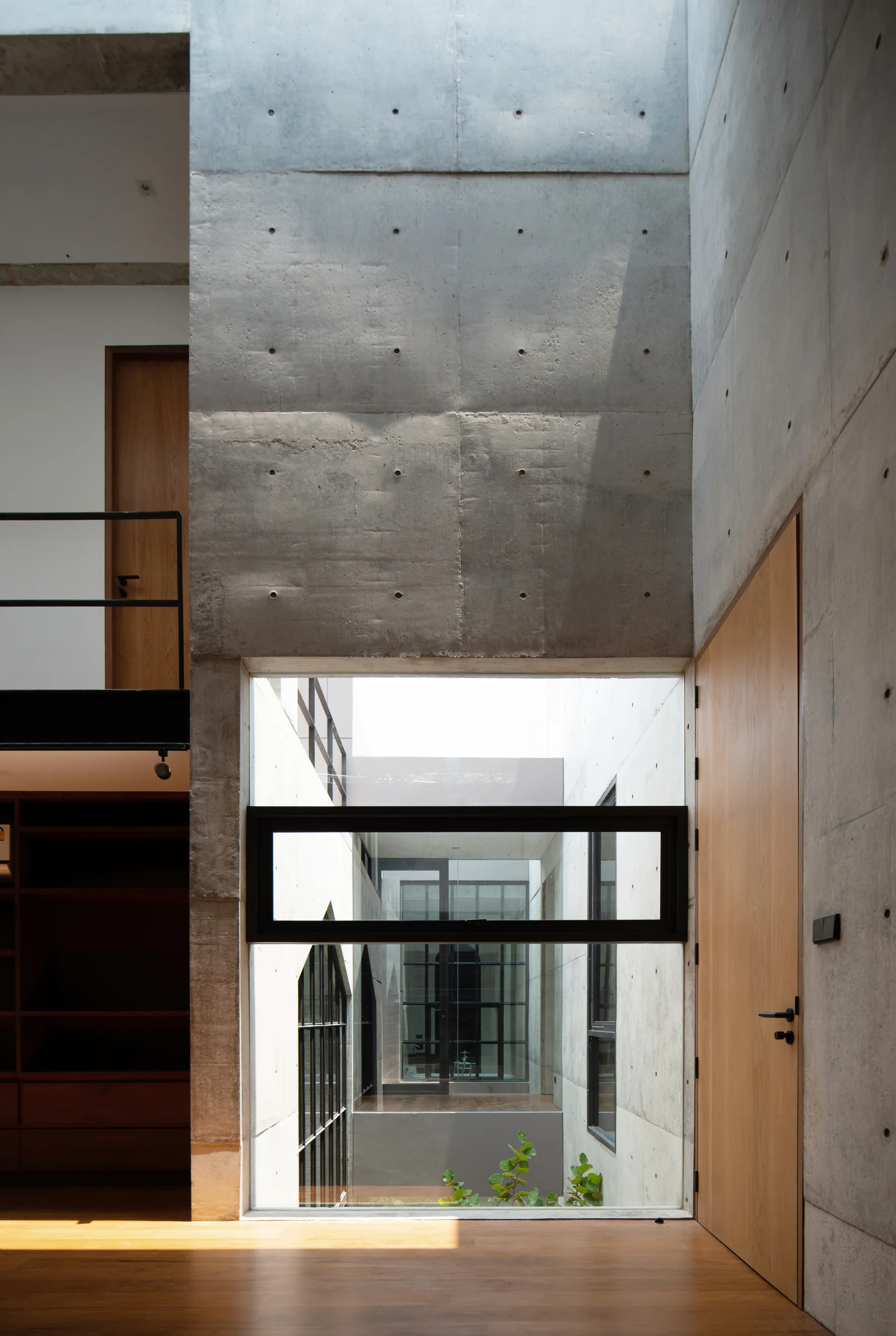 A modern interior hallway featuring raw board-formed concrete walls, warm wood doors, and a window viewing an inner lightwell.