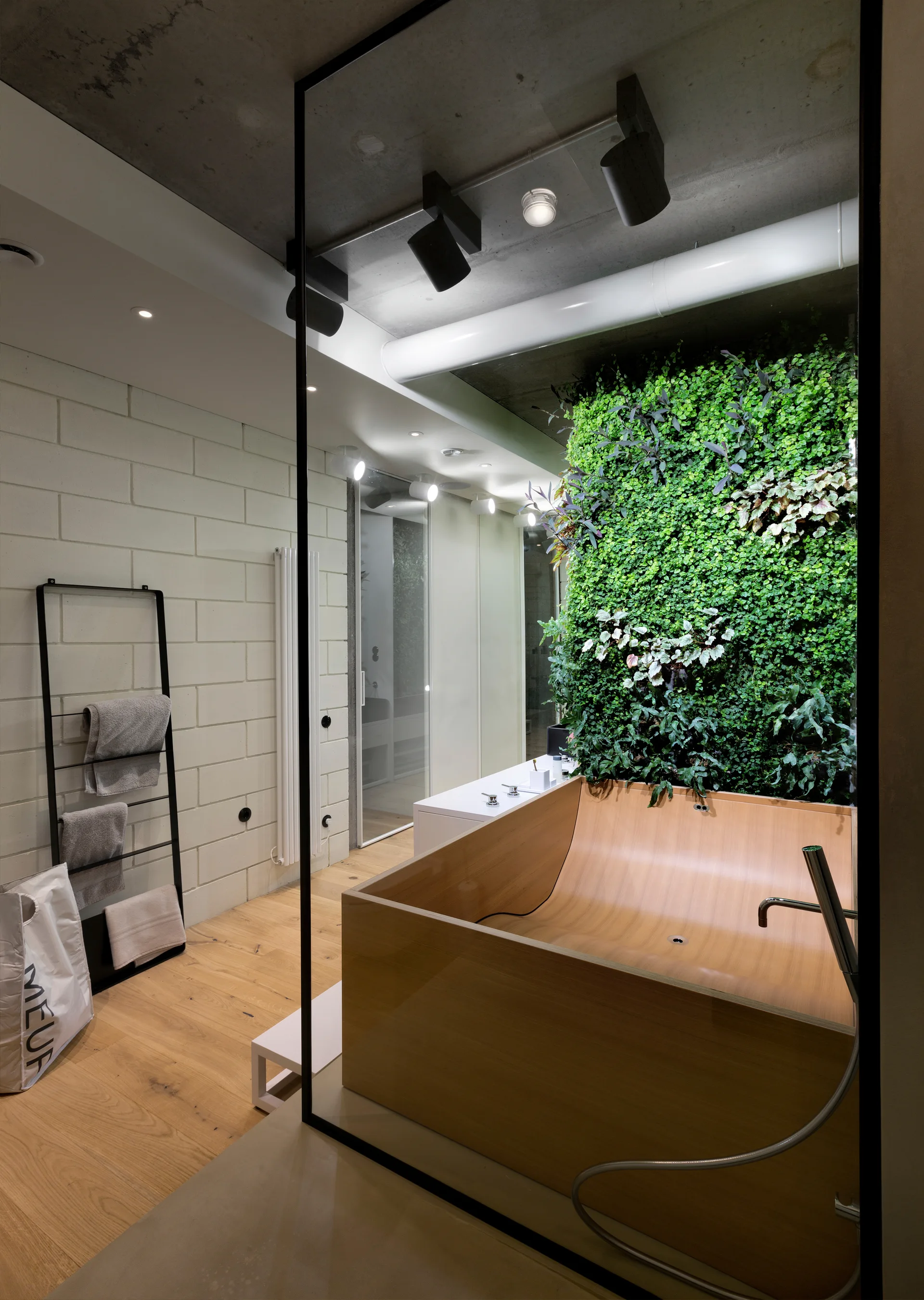 A modern bathroom features a square wooden bathtub set against a vertical living green wall, with an exposed concrete ceiling above.