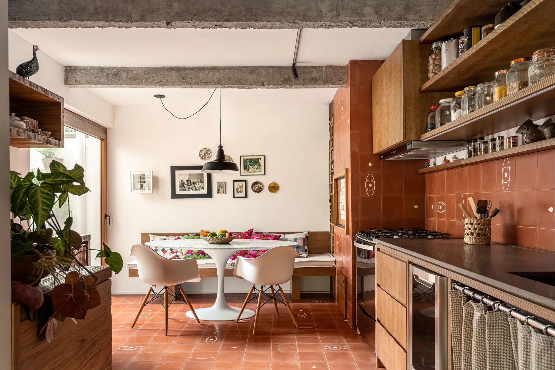 A kitchen and dining area features terracotta tile flooring and walls, a built-in wooden bench, a round white tulip table, and exposed concrete ceiling beams.