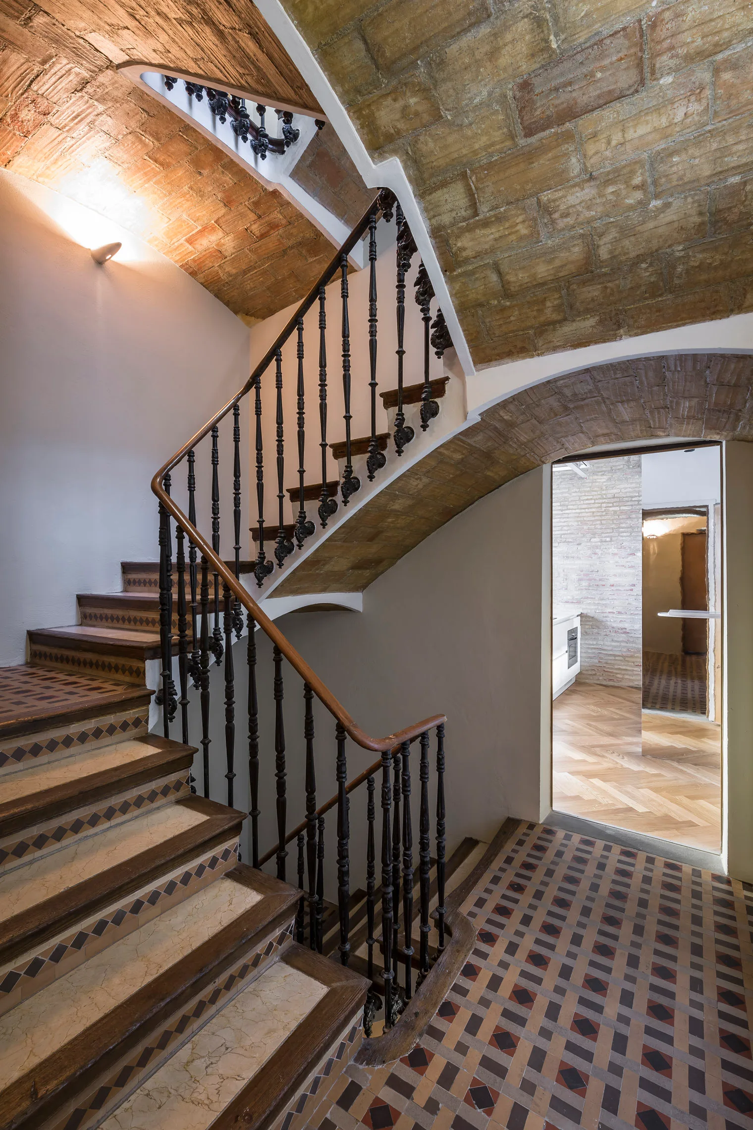 A multi-level staircase features patterned tile risers, dark metal railings, and a wooden handrail beneath a vaulted brick ceiling.