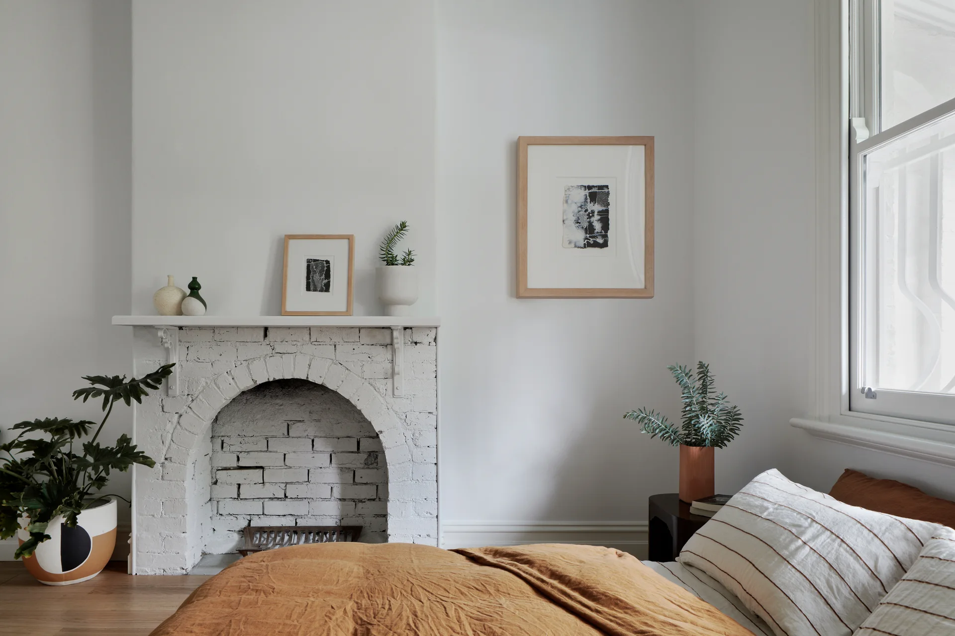 A minimalist bedroom features a white painted brick fireplace, a bed with warm ochre linen bedding, striped pillows, and simple wood-framed wall art.