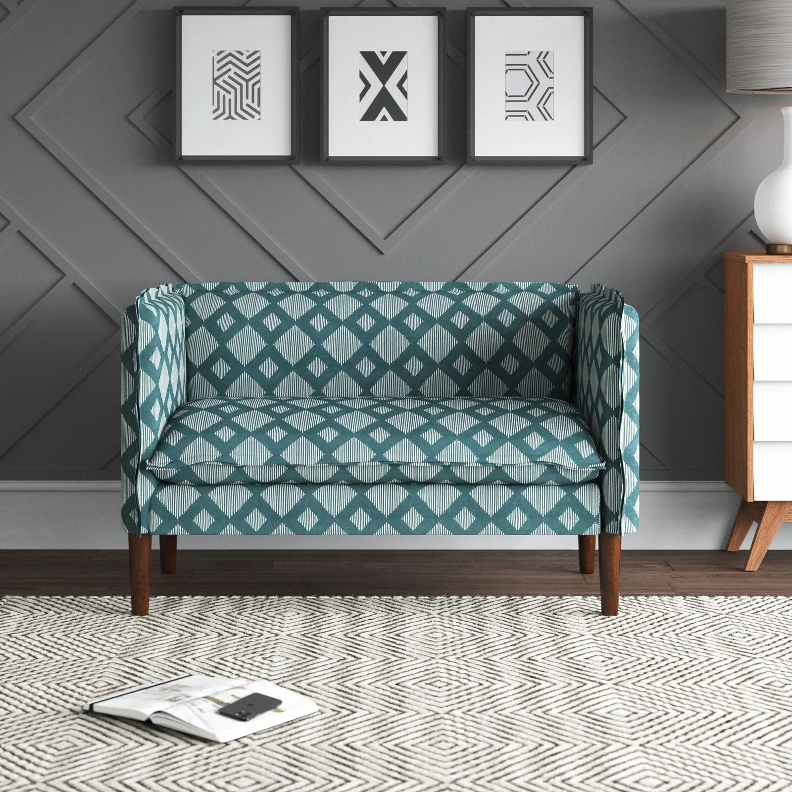 A teal patterned loveseat sits against a grey paneled wall featuring three framed prints, placed on a patterned rug next to a wooden chest.