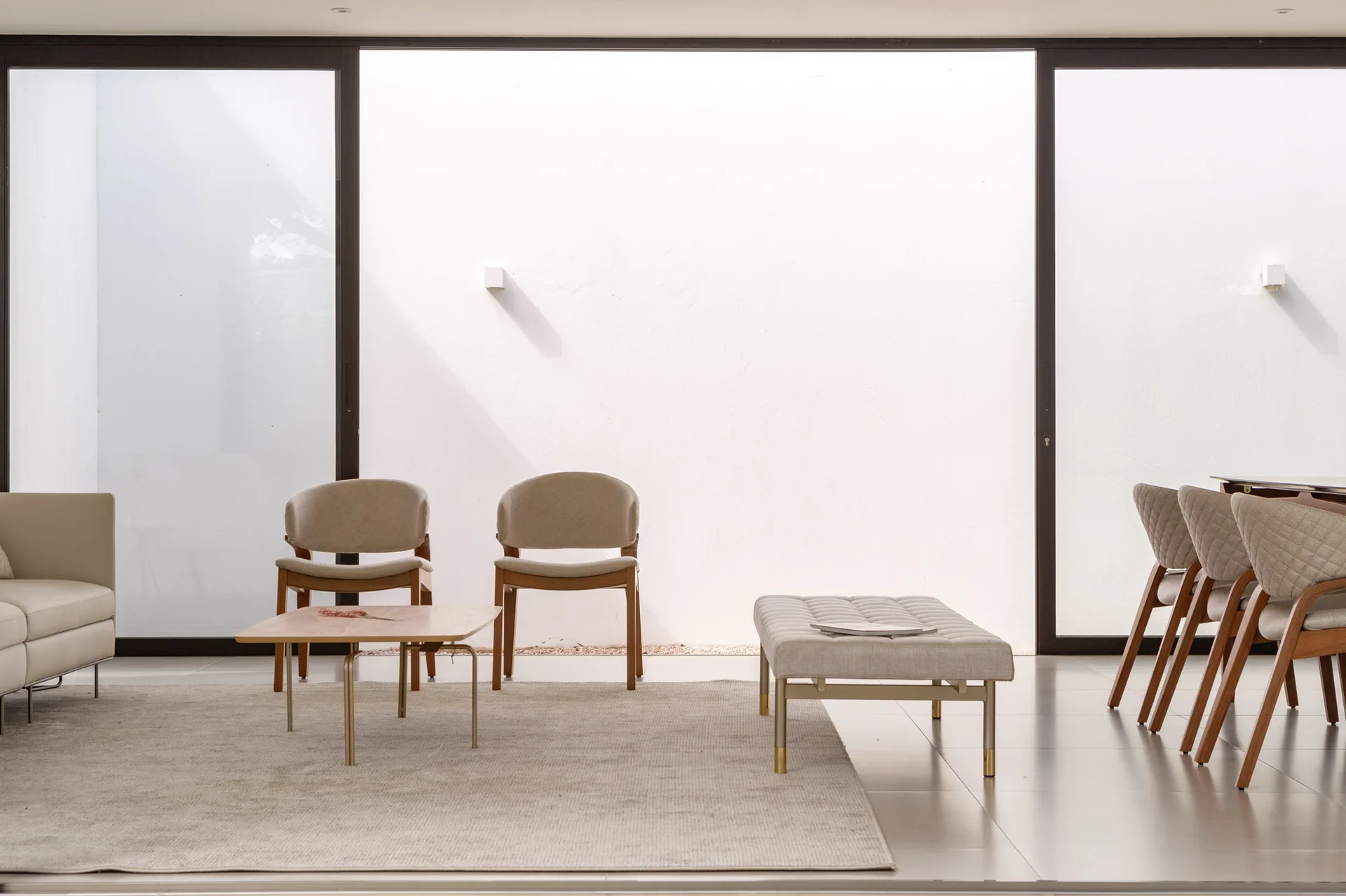 A minimalist living room featuring a pair of beige upholstered wood chairs, a tufted bench, and a low coffee table in front of large sliding glass doors.
