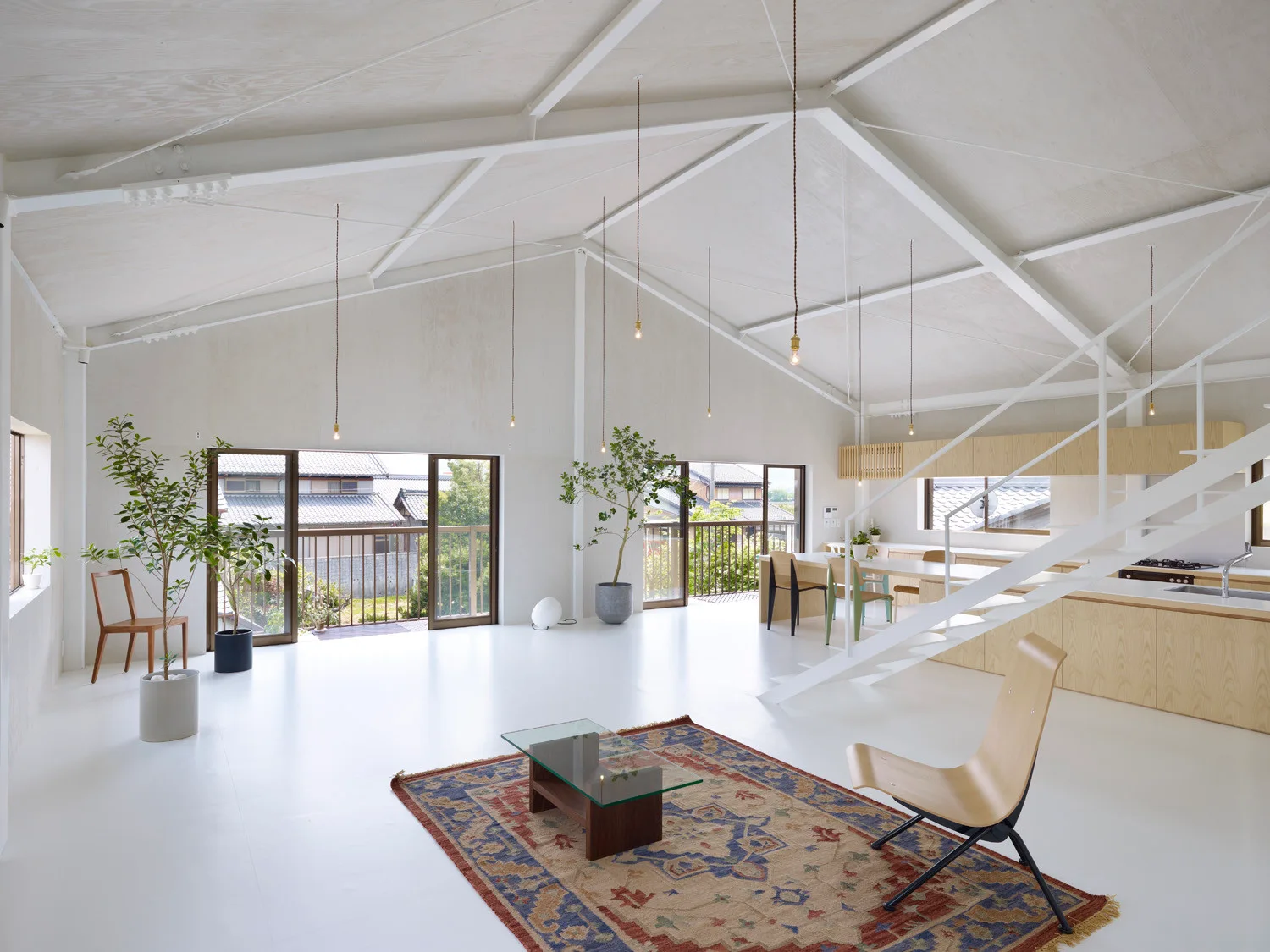 An airy open-plan living and kitchen space featuring white floors, exposed ceiling rafters, a white steel staircase, and a patterned area rug.